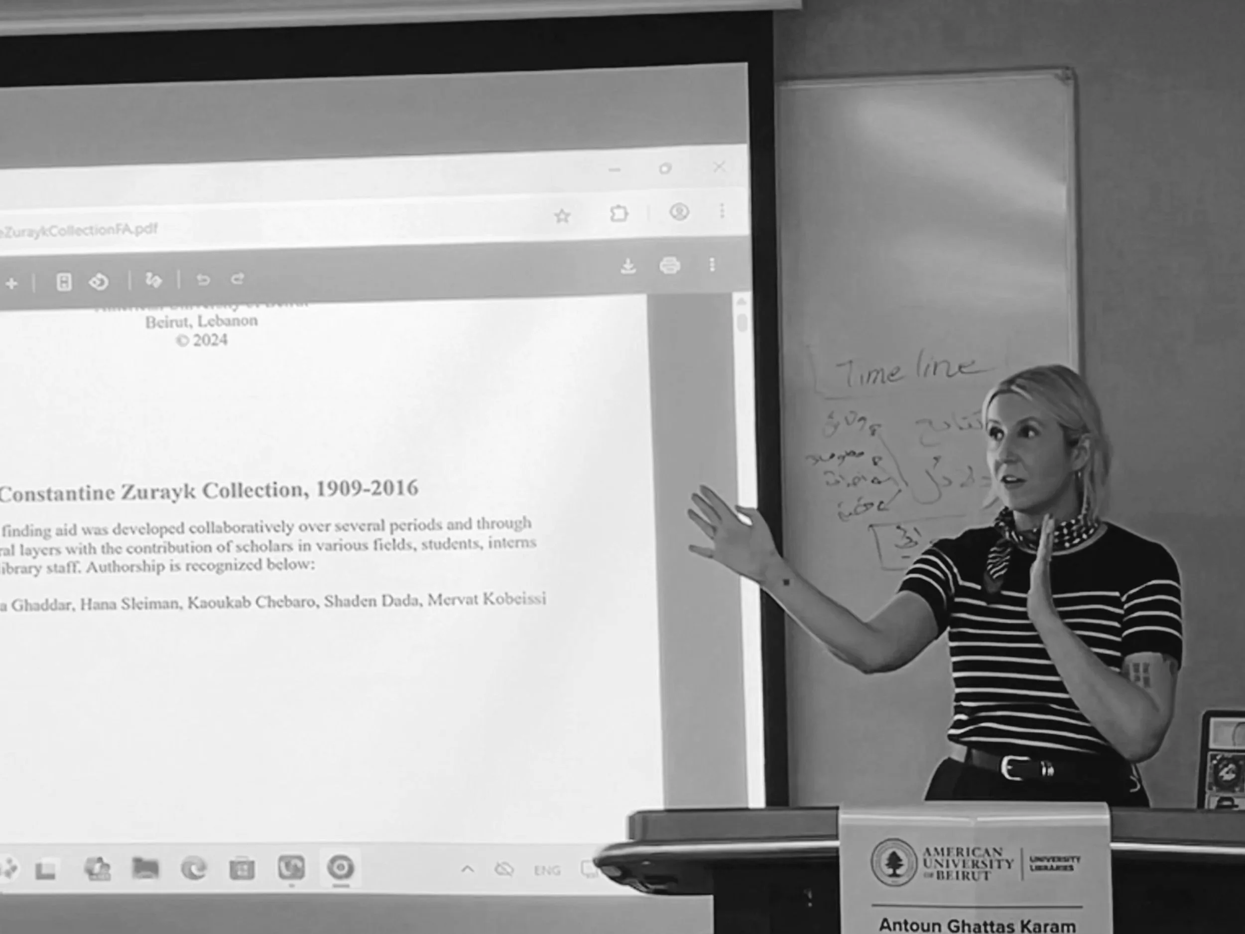A woman standing at a podium in front of a classroom, gesturing with her right hand while pointing at a projected document on a whiteboard behind her. The document is titled "Constantine Zurayk Collection, 1909-2016" and contains text and author names. She has blonde hair, wears a striped shirt, and a polka-dot scarf. There is a label on the podium reading "Antoun Ghattas Karam," and the whiteboard has some handwritten notes including "Timeline" in English and other text in Arabic.