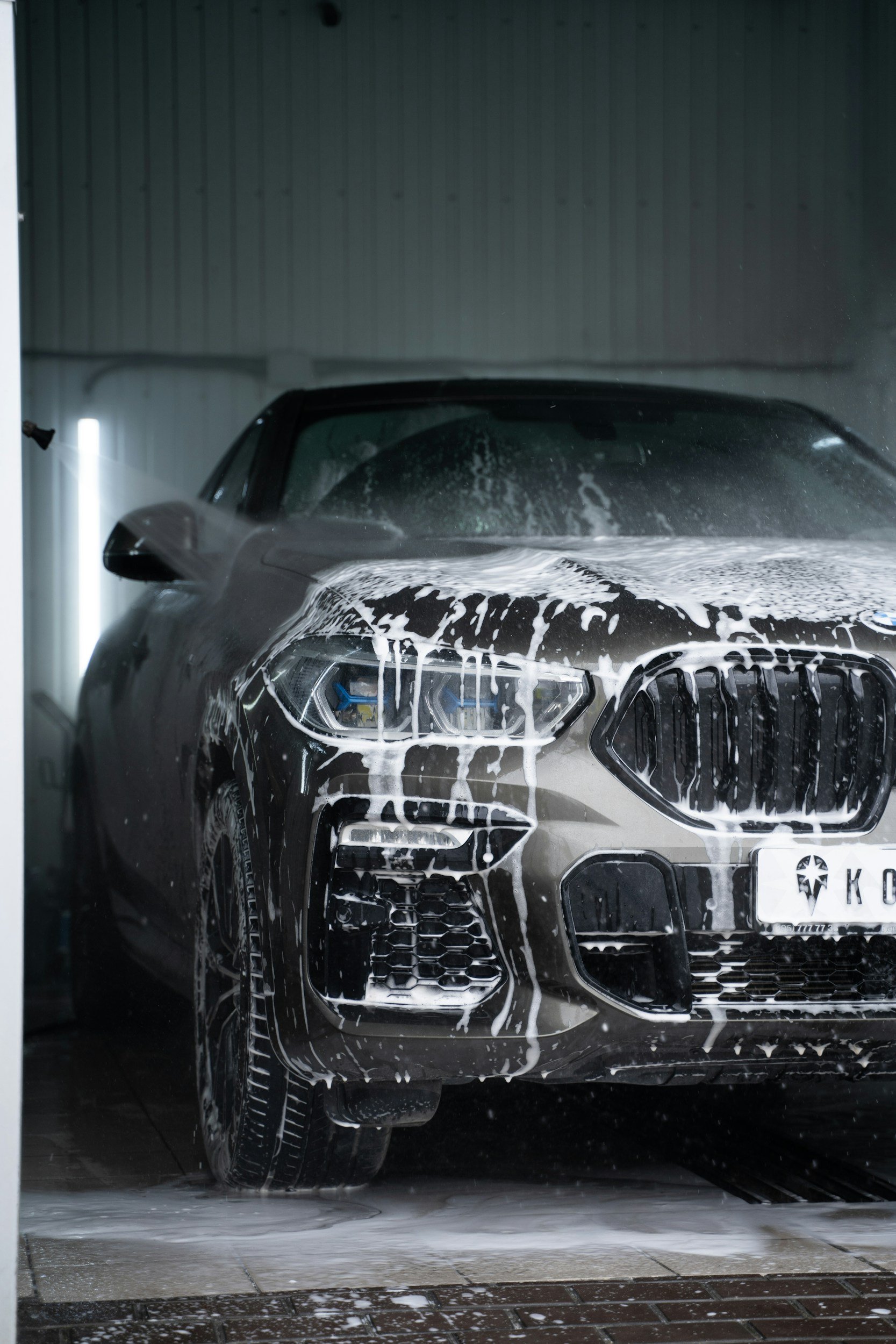 A dark-colored luxury car, possibly an Audi, being washed with soap in a car wash bay. The foam covers the front of the car, including the hood, front grill, and headlights.