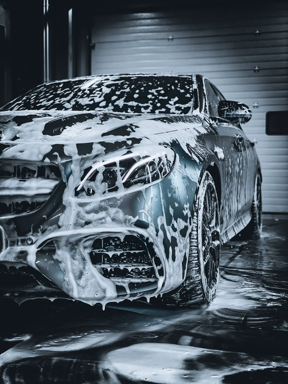A black sports car being washed with soap and water, inside a garage with a closed roll-up door.