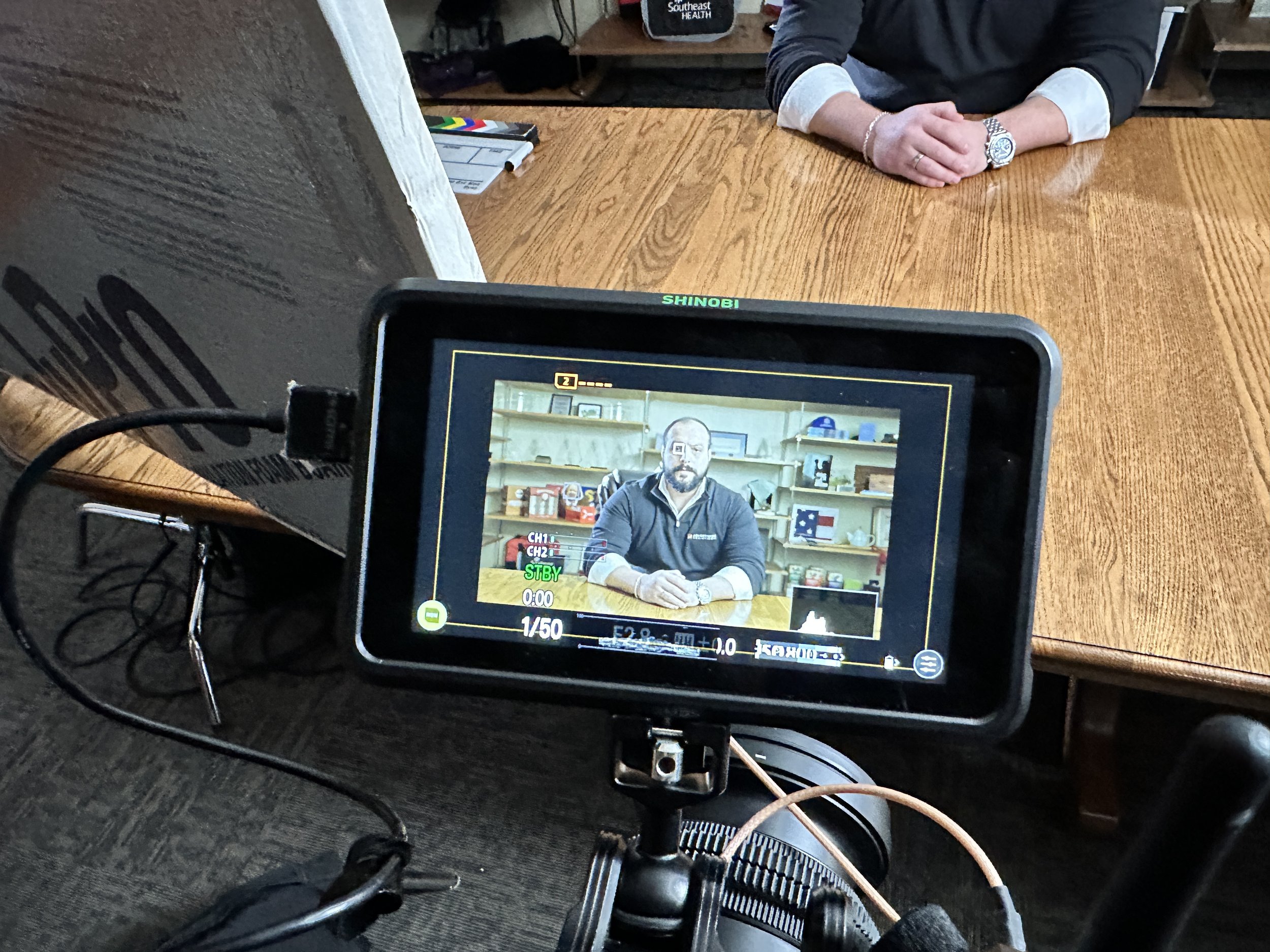 A camera monitor shows a man sitting at a wooden table with children and adult supplies in the background. The man has a beard and is wearing a dark jacket, and is seated in a room with shelves containing various items.
