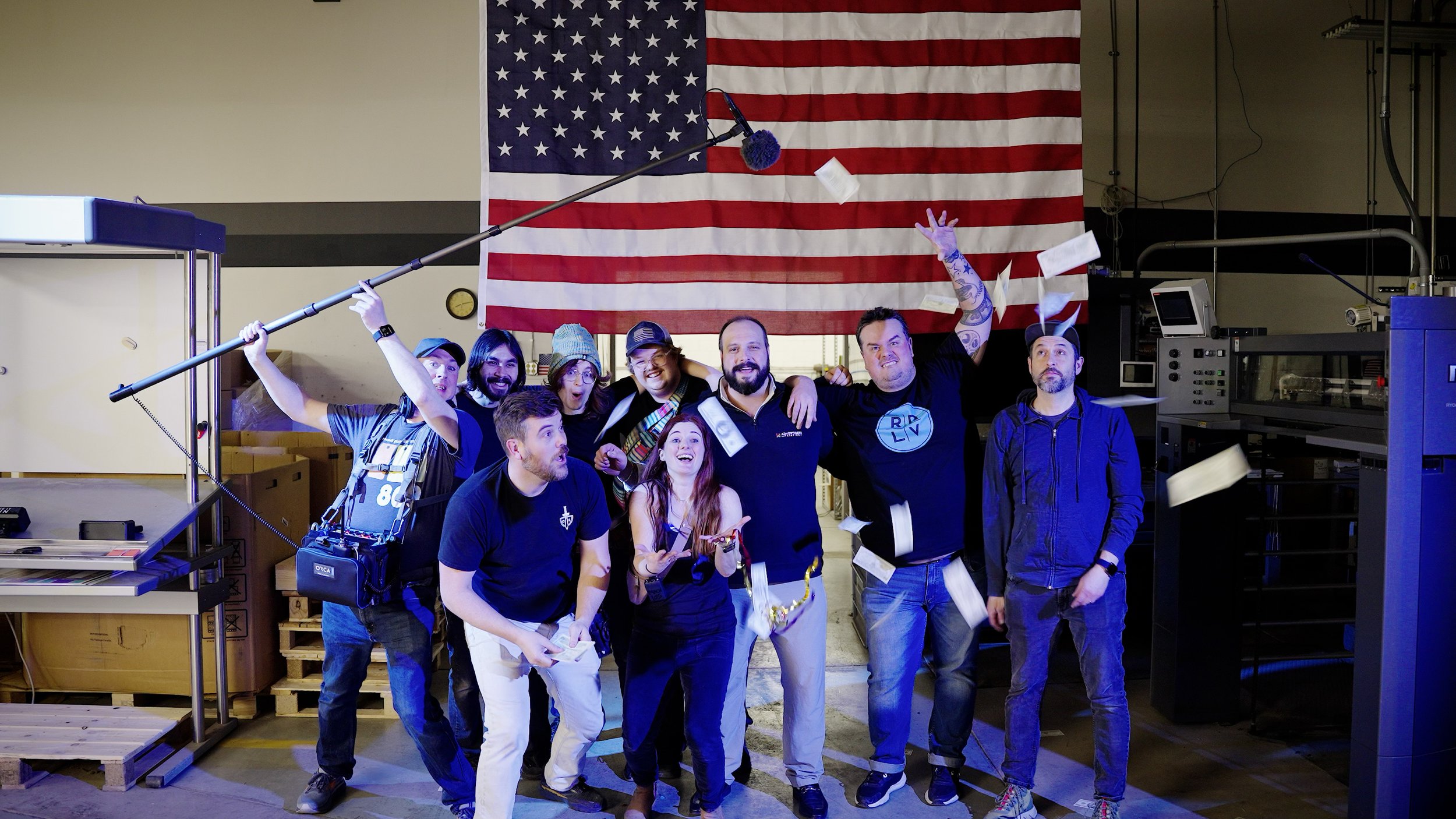 Group of eight people celebrating with a large American flag in the background, confetti flying in the air, some smiling and one holding a boom microphone, in an indoor setting.