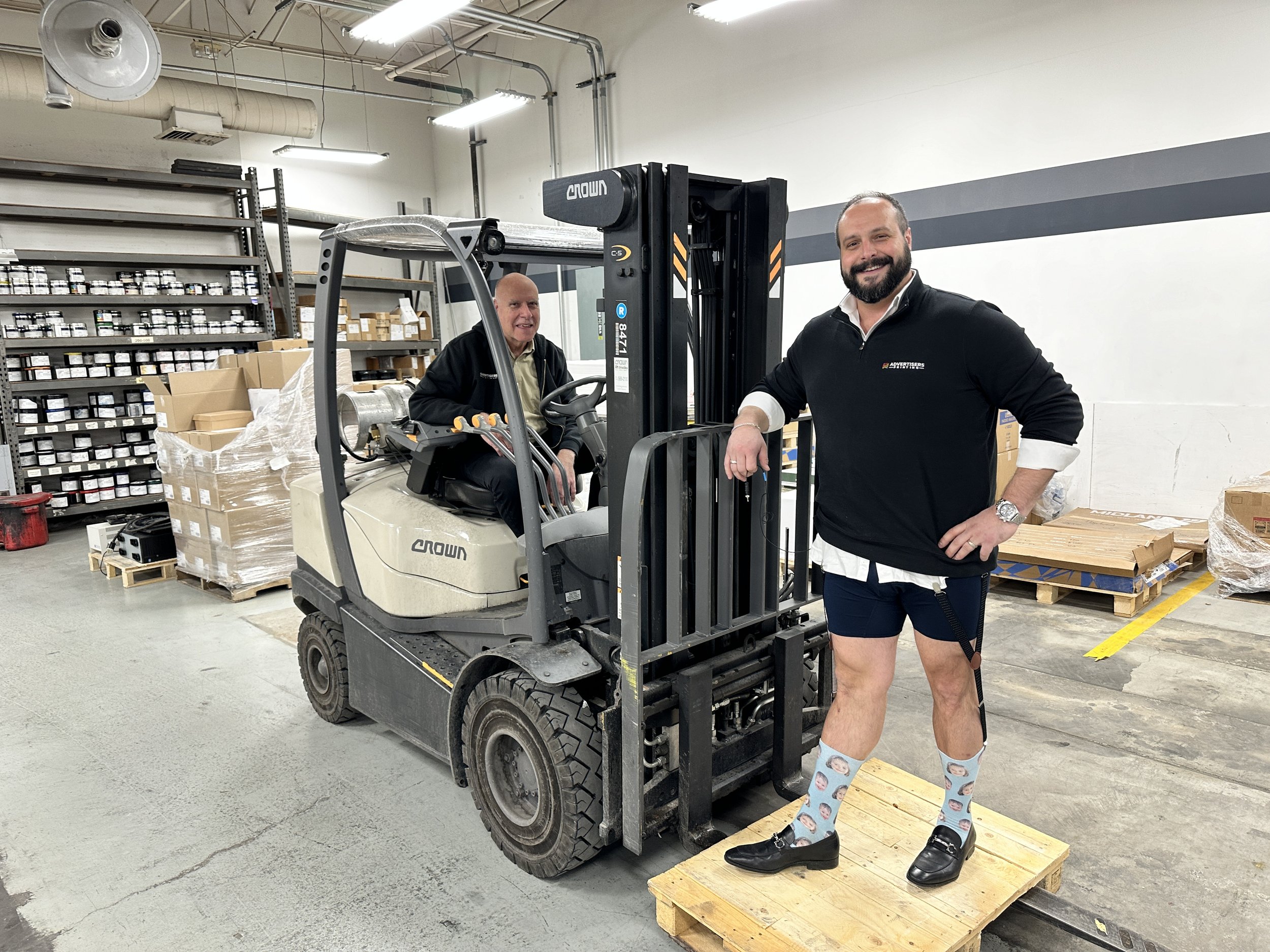 Two men in a warehouse, one sitting on a forklift and the other standing on a wooden pallet, posing for the photo. The man on the pallet is wearing funny socks and dress shoes, and the forklift operator is sitting on the forklift.