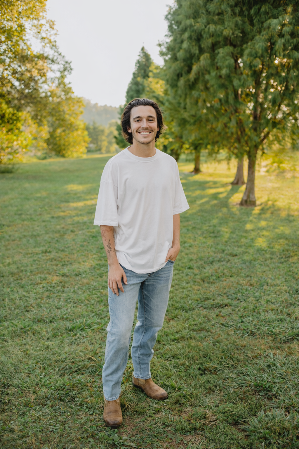 A young man with dark hair, facial hair, wearing a white oversized t-shirt, light blue jeans, and brown boots standing on a grassy field in a park with green trees in the background, smiling at the camera.