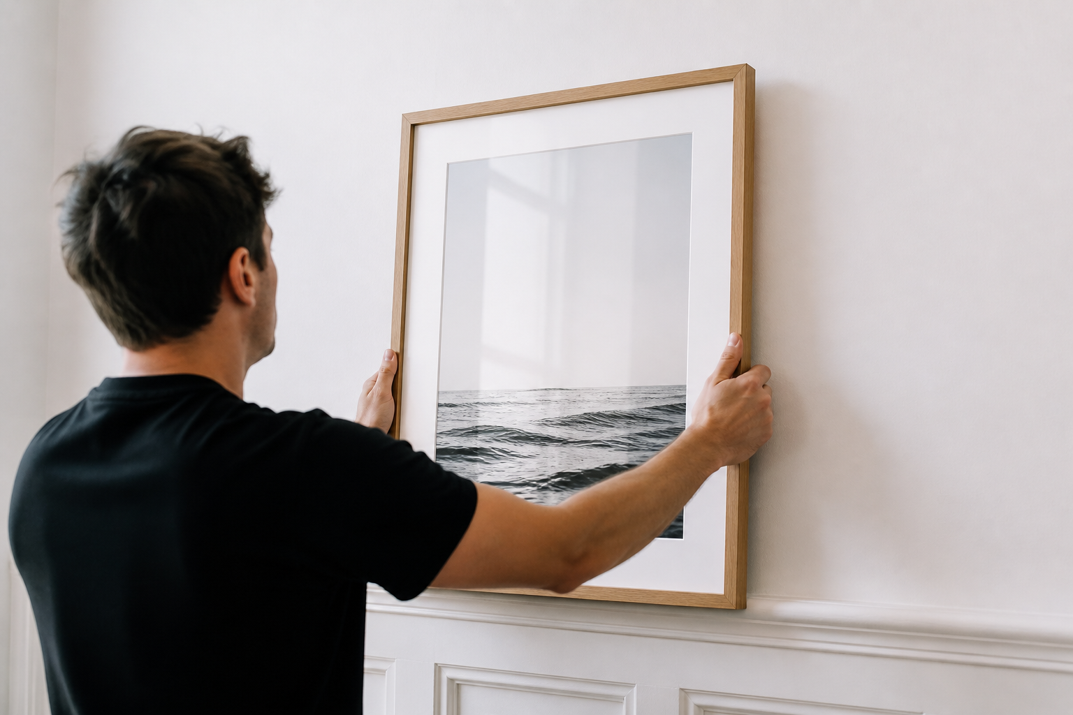 Person hanging framed black and white ocean photograph on a white wall.