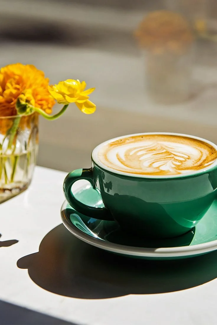A green cup of coffee with latte art on top, placed on a matching saucer, and a glass vase with yellow and orange flowers in the background.
