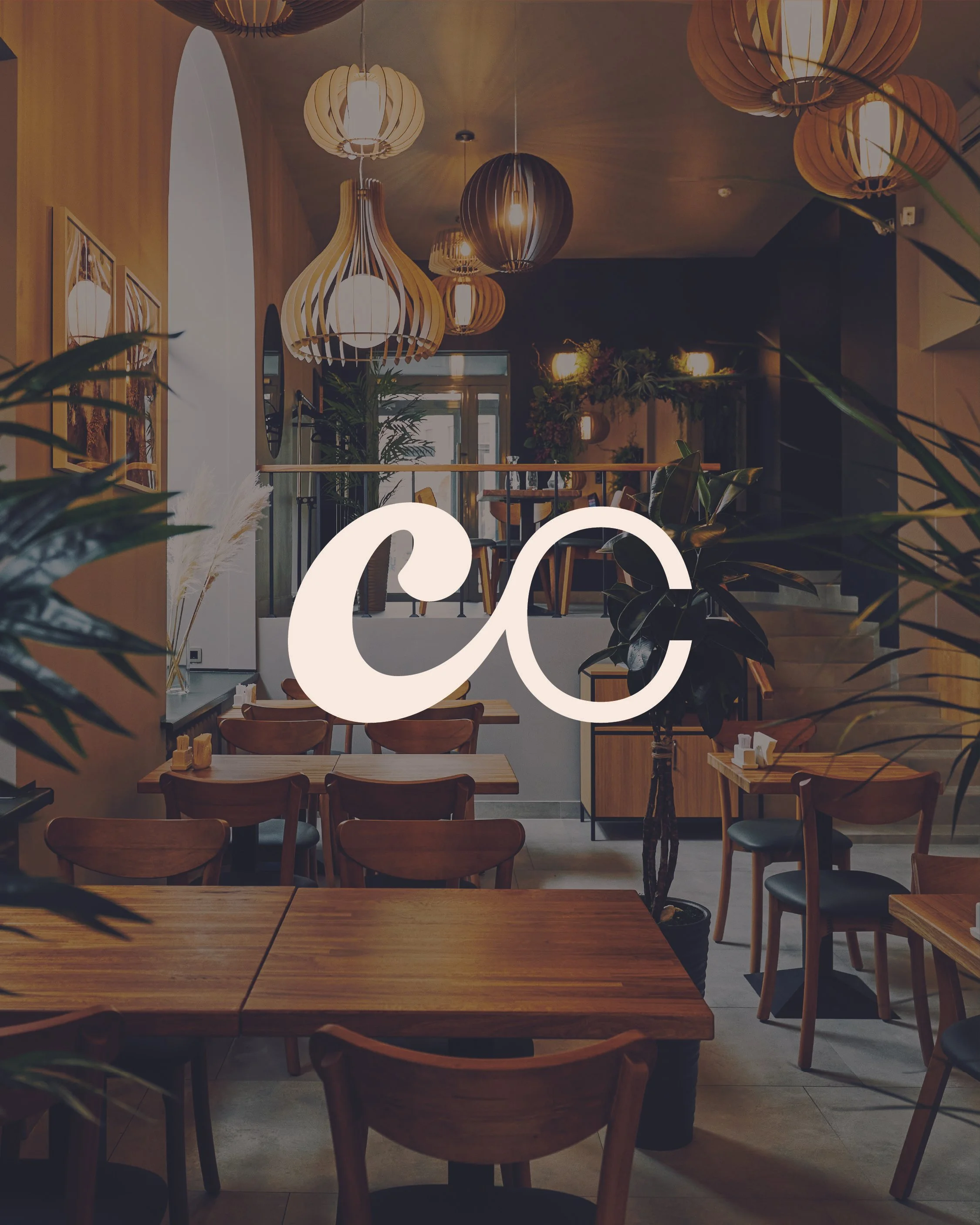 Interior of a cozy restaurant with wooden tables and chairs, hanging pendant lights, and green plants, viewed through a glass window with a white logo.