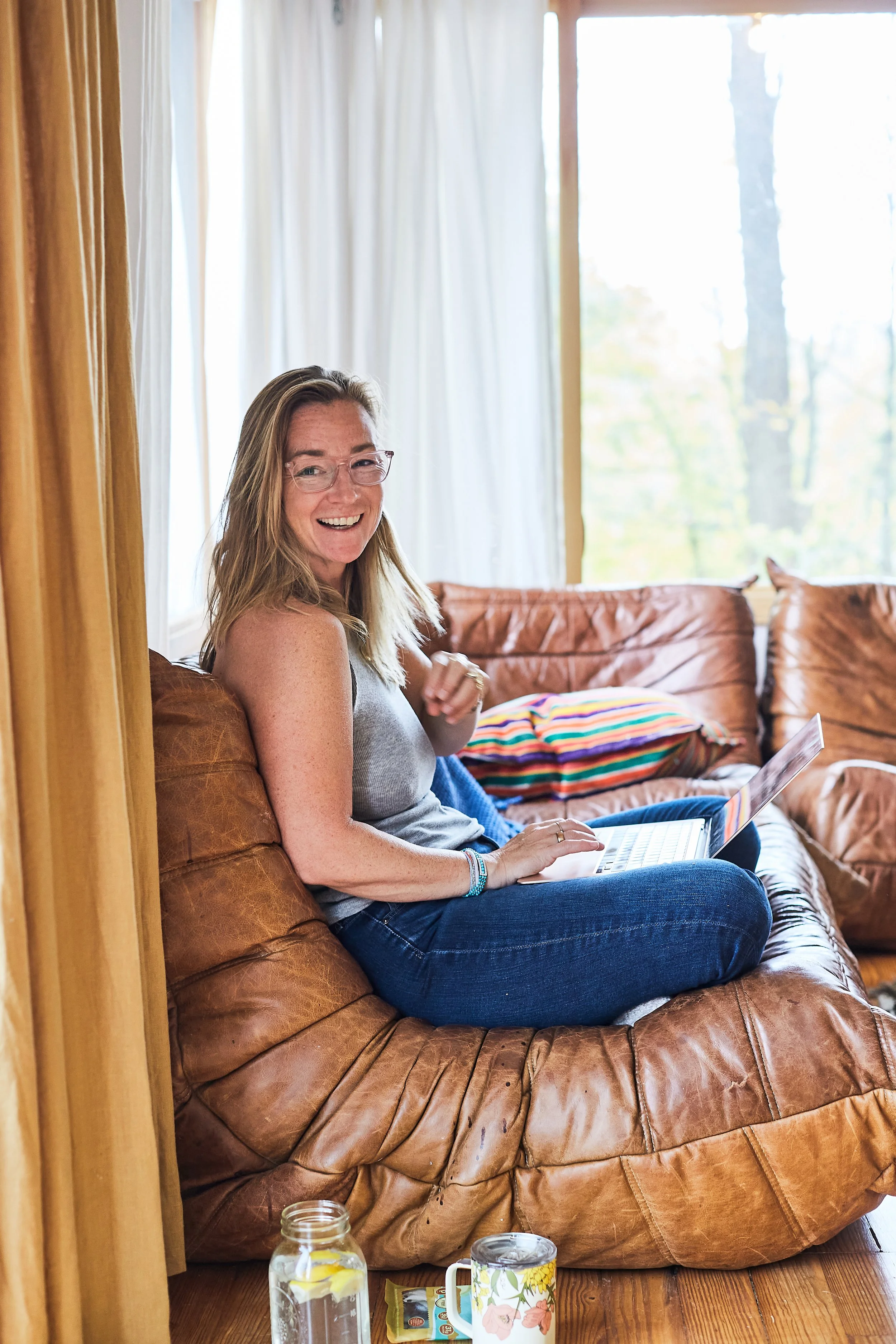 A woman with glasses sitting on a leather couch using a laptop, smiling, with a jar of water and a mug on the floor beside her in a cozy, well-lit room.