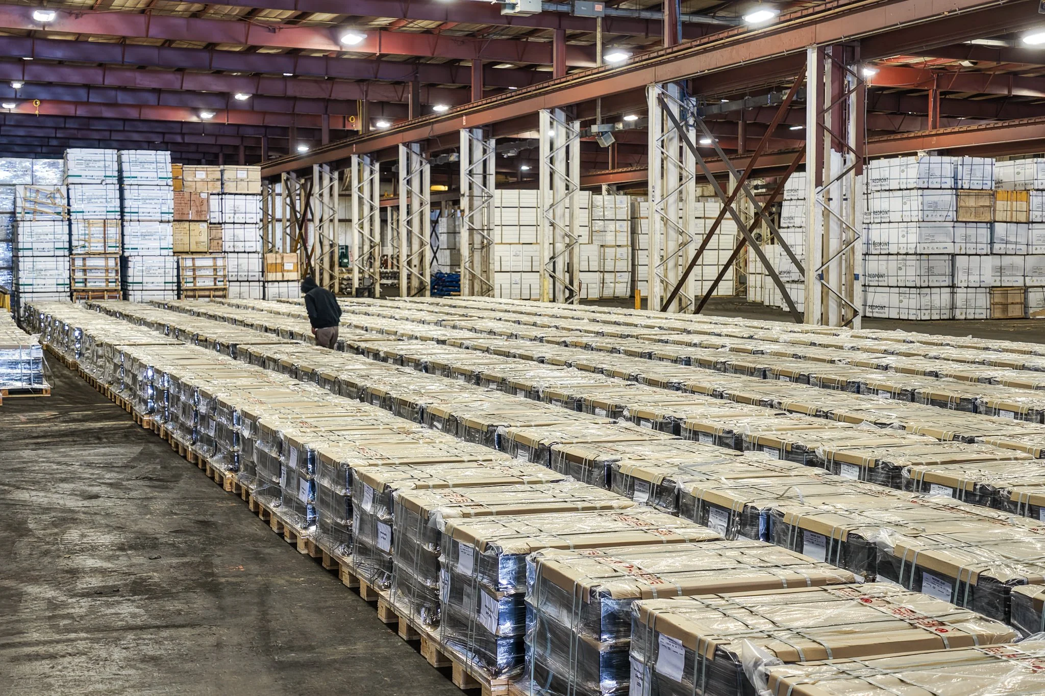 A warehouse filled with large stacks of boxed products arranged on pallets, with a person inspecting the boxes.