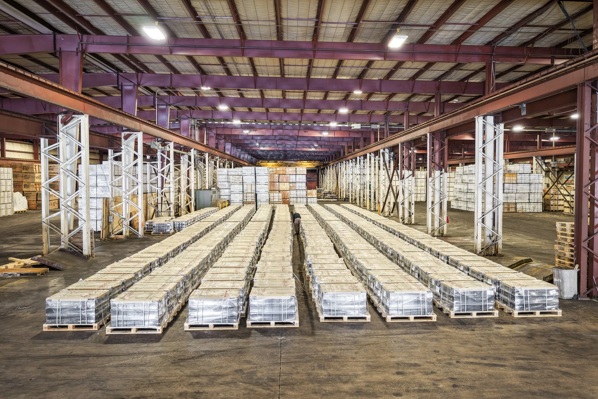 Stacks of cardboard boxes wrapped in plastic on wooden pallets inside a warehouse with metal beams and a high ceiling.