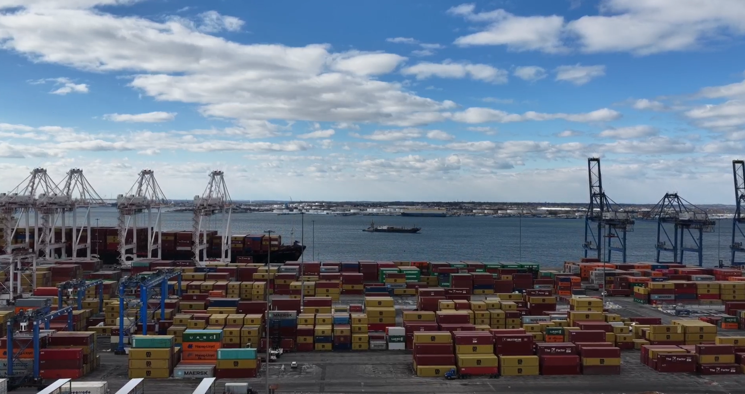 Port with shipping containers, cranes, and ships on the water, under a partly cloudy sky.