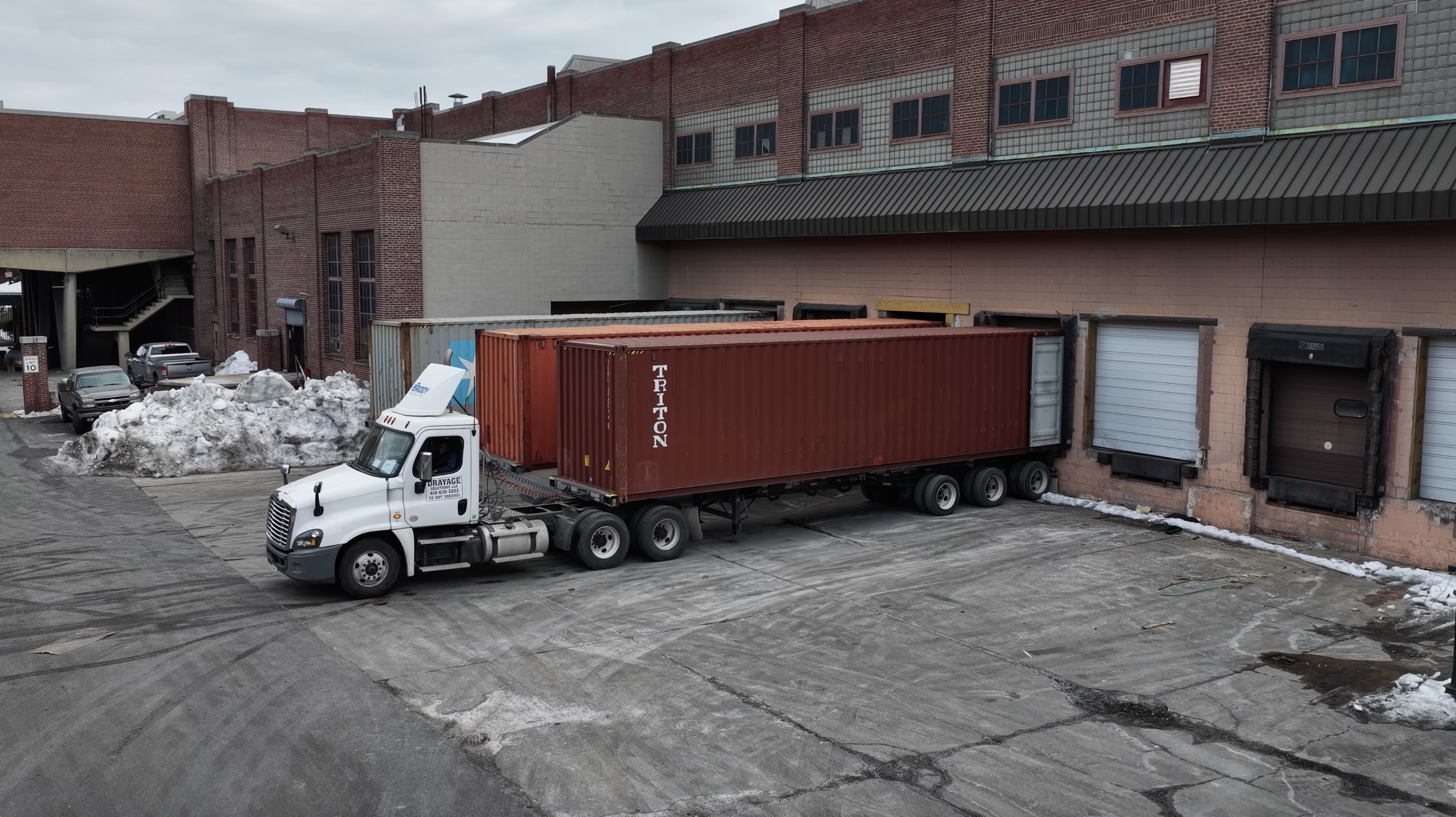 A white semi-truck with a red shipping container parked next to a brick building with loading docks, in a parking lot with snow piles and a few parked cars, on an overcast day.