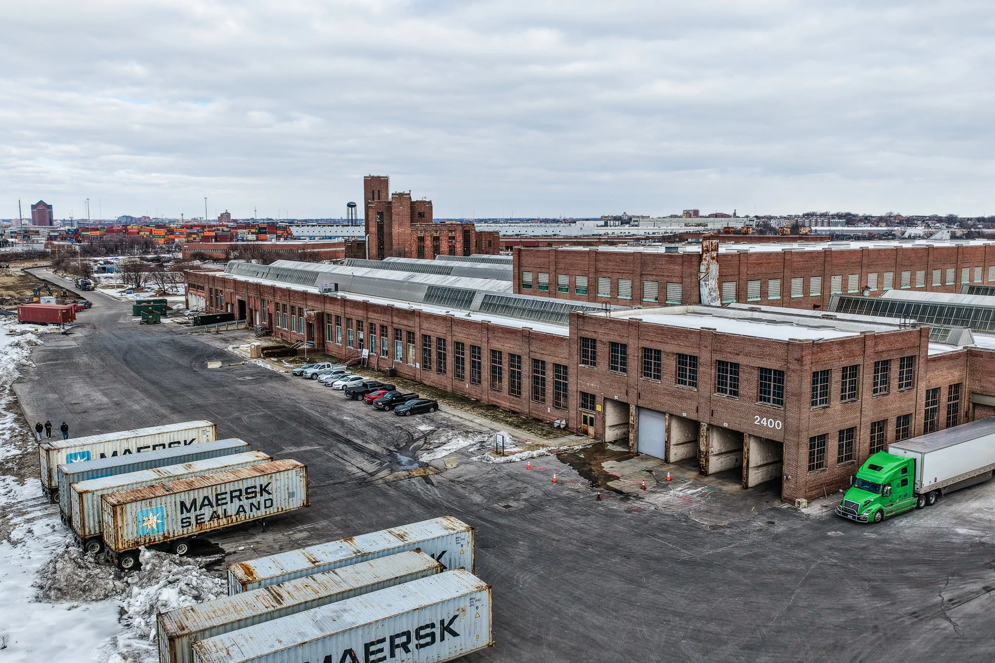 An industrial warehouse with red brick exterior, large windows, and a parking lot. Shipping containers are in the foreground, with snow on the ground. A green semi-truck is parked near the building's entrance. Overcast sky above.