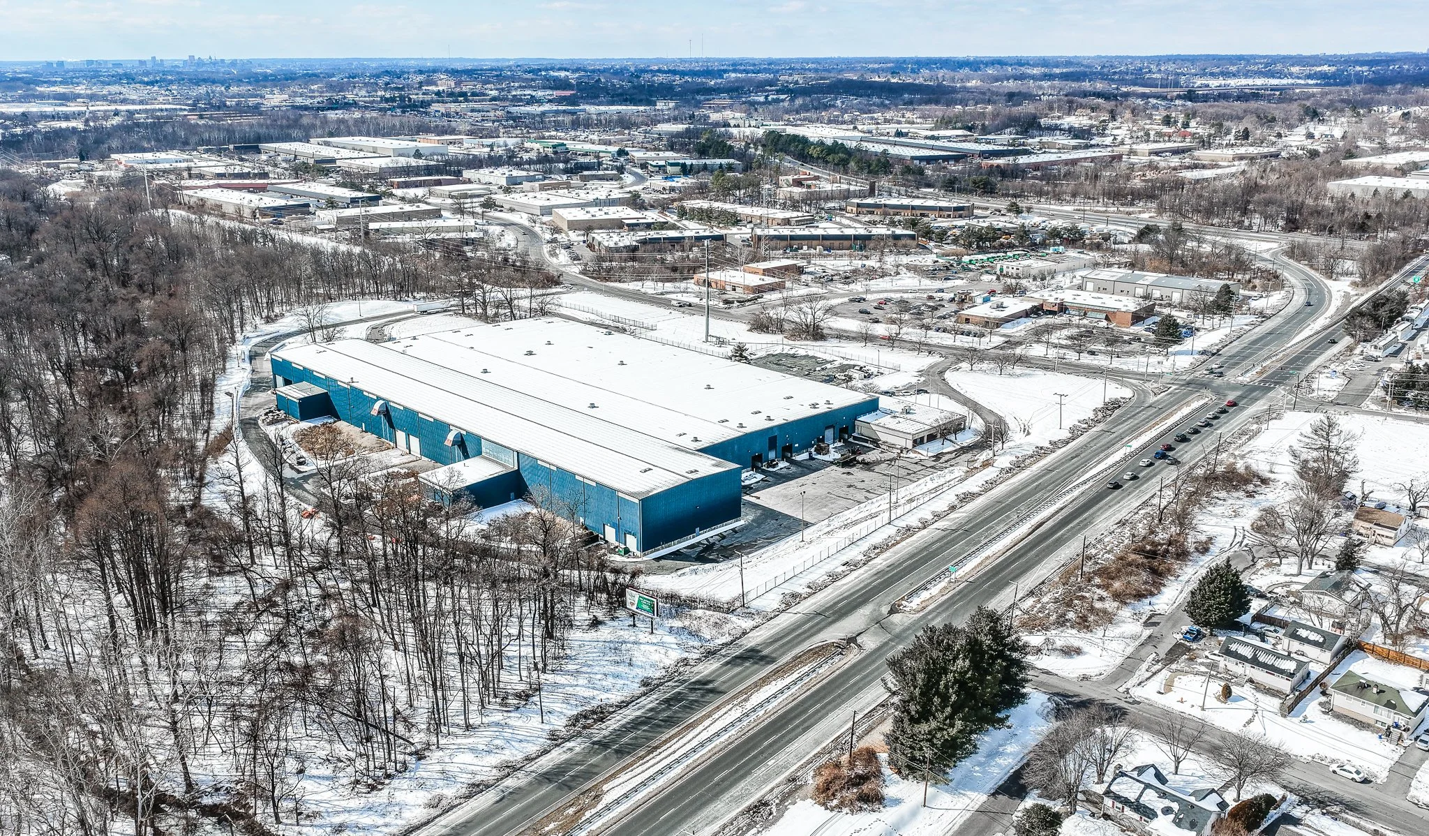 Aerial view of a snowy industrial area with a large blue warehouse, surrounding roads, and snow-covered trees and residential houses.