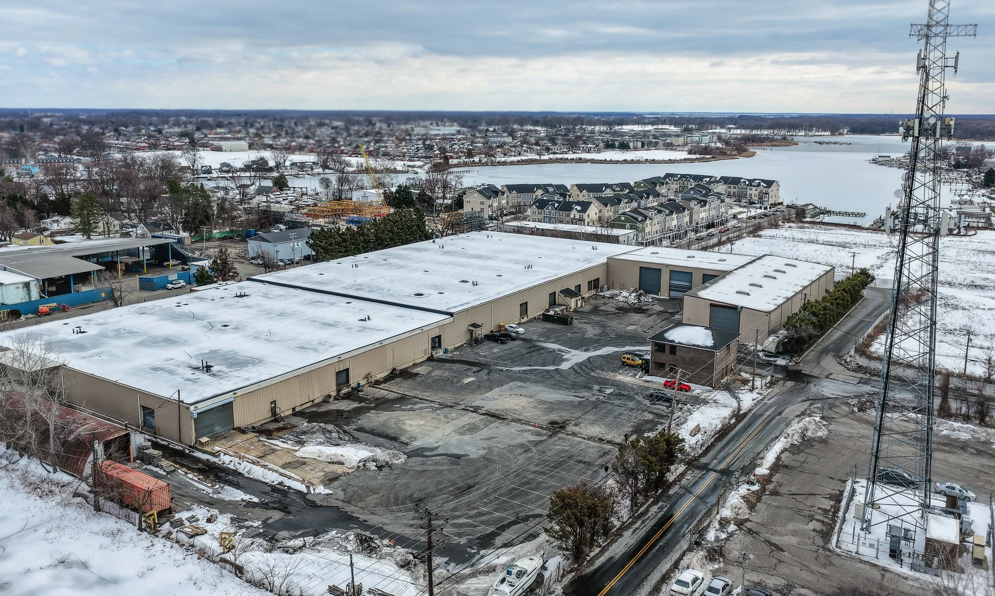 Aerial view of a snow-covered industrial building with parking lot, surrounded by snowy landscape and residential area, with a river in the background and a tall telecommunications tower on the right.