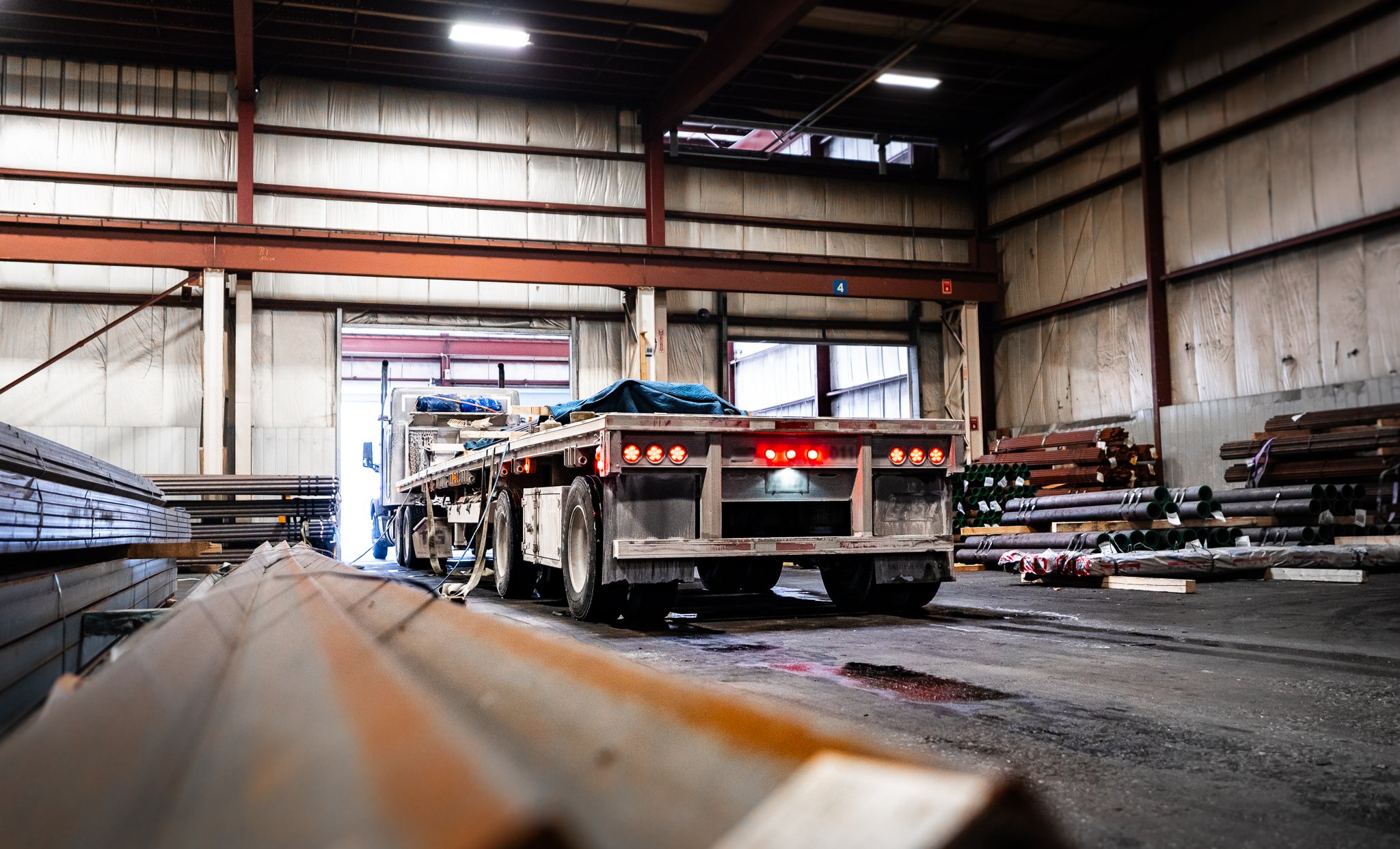 Inside an industrial warehouse with a flatbed truck parked near the open garage door, surrounded by stacks of metal pipes and construction materials.