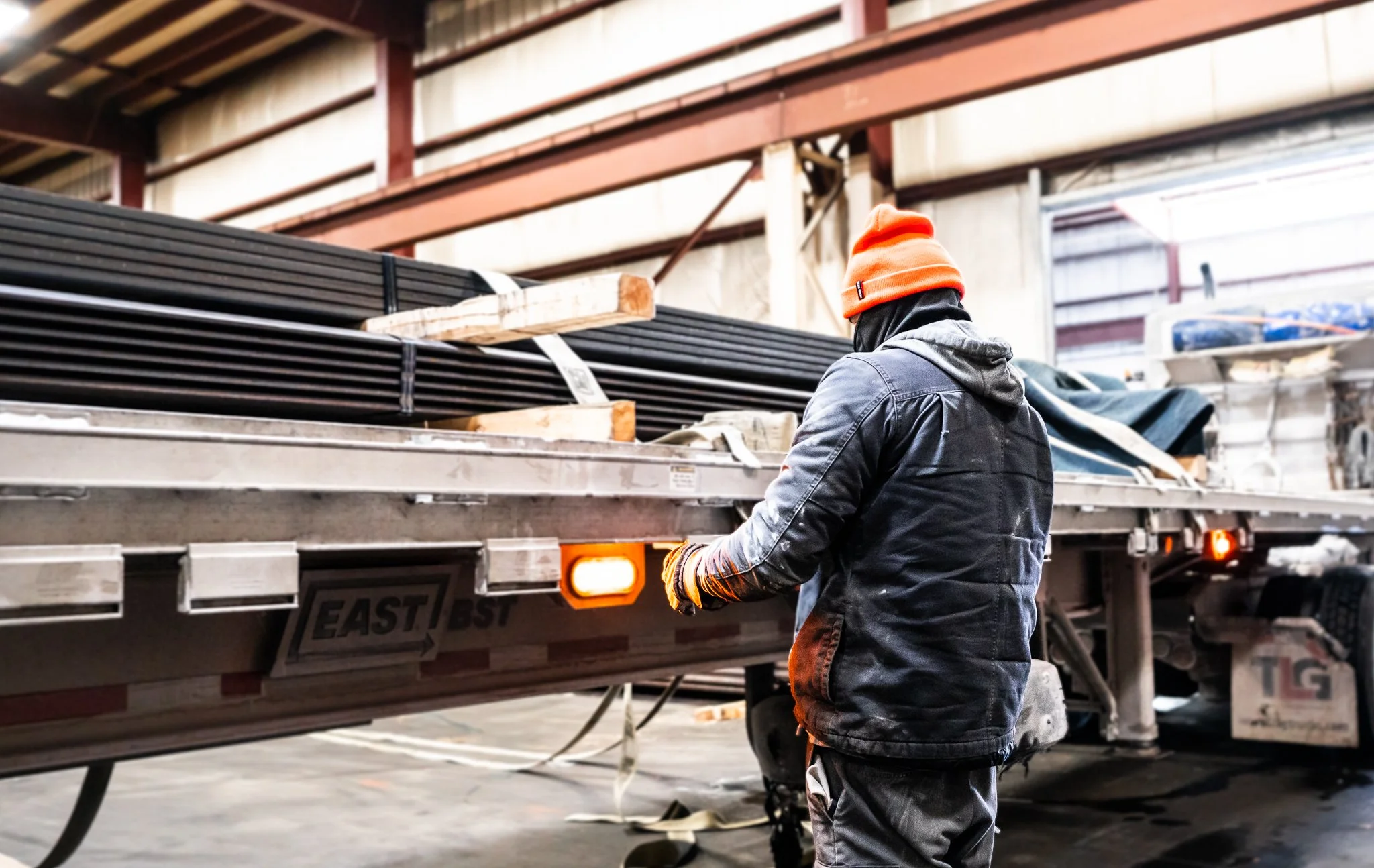 A person wearing a black jacket and orange beanie working near a truck in an industrial warehouse.