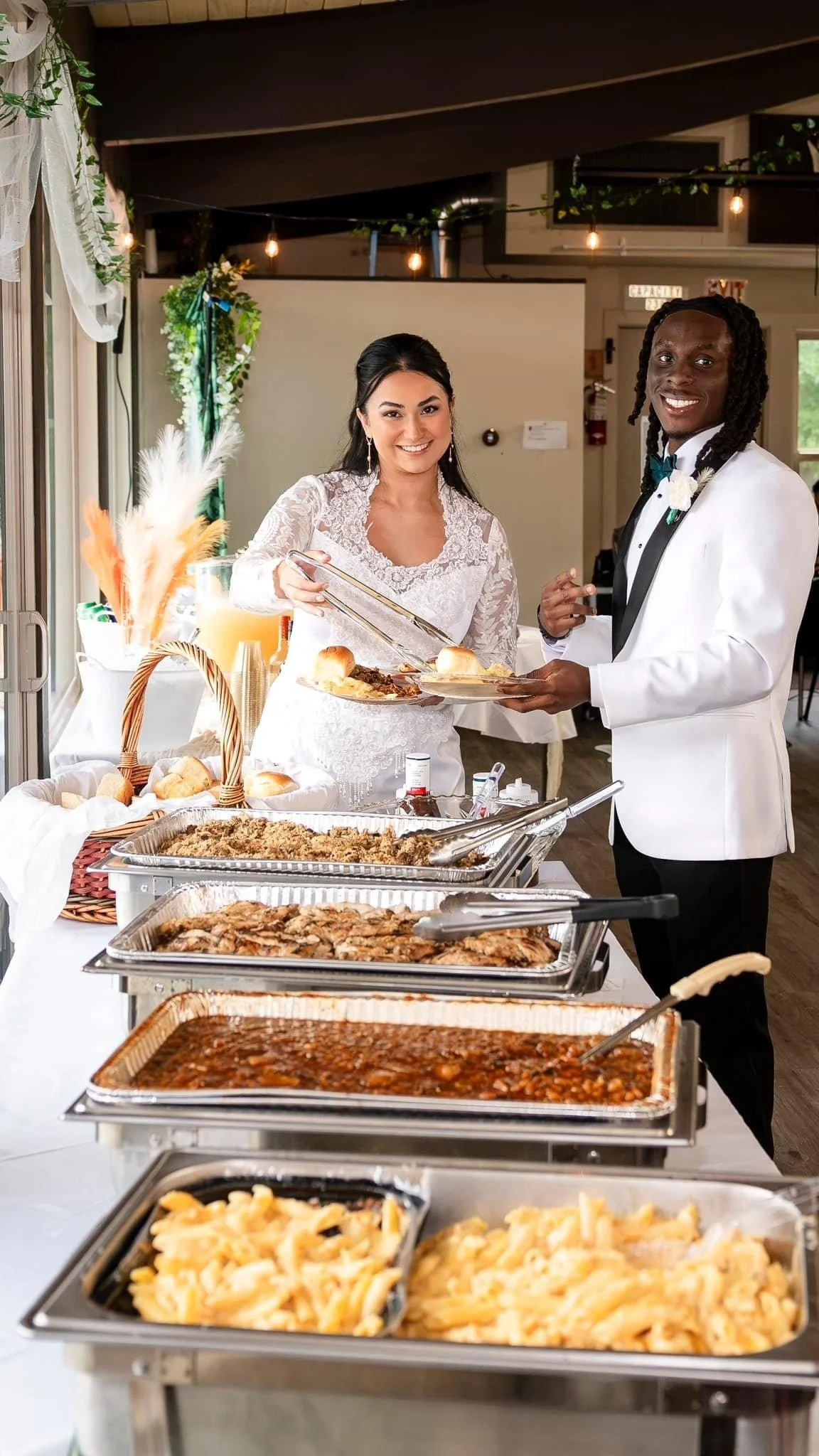A bride and groom serve themselves food at a wedding reception buffet table with various dishes like pasta, chili, chicken, and bread, inside a decorated venue with string lights.