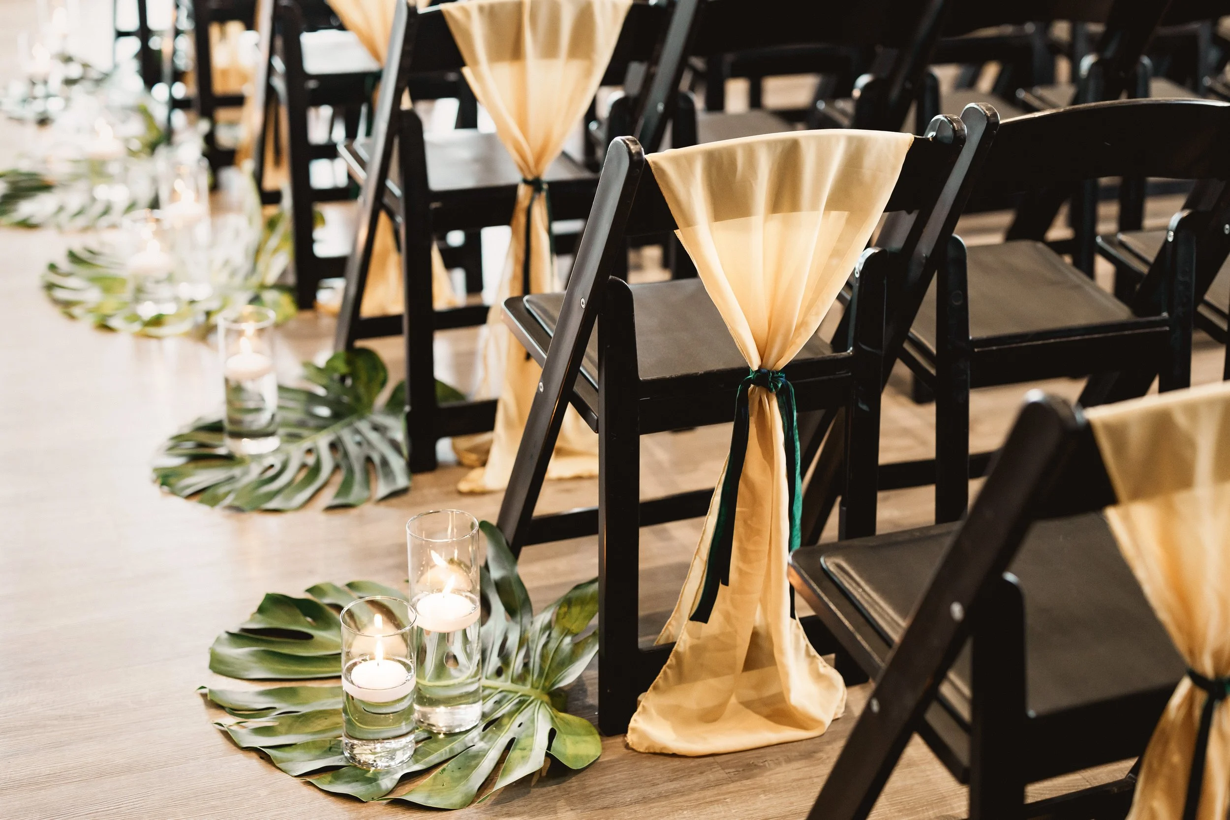 Black ceremony chairs are layered with gold fabric sash ties on the backs, the aisle lined with tropical green leaves and tea lights.