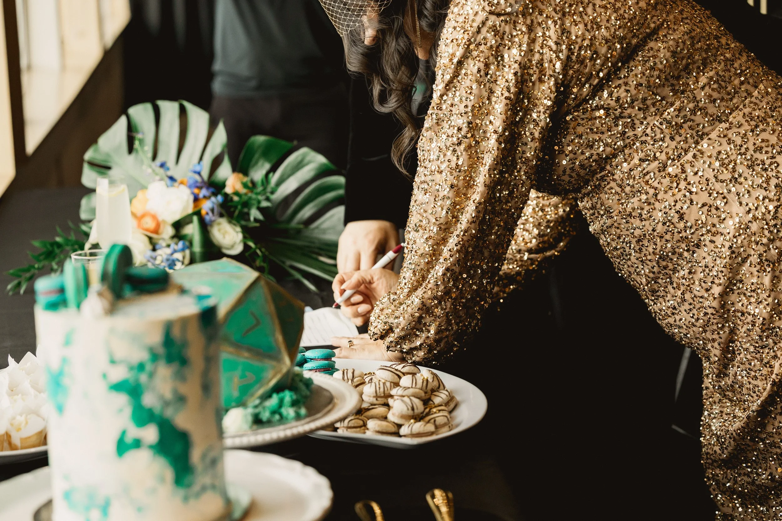 The bride and groom in gold sequins and black velvet are signing their marriage licence at a decorated table with a selection of cakes, desserts, flowers, and a large tropical floral arrangement.