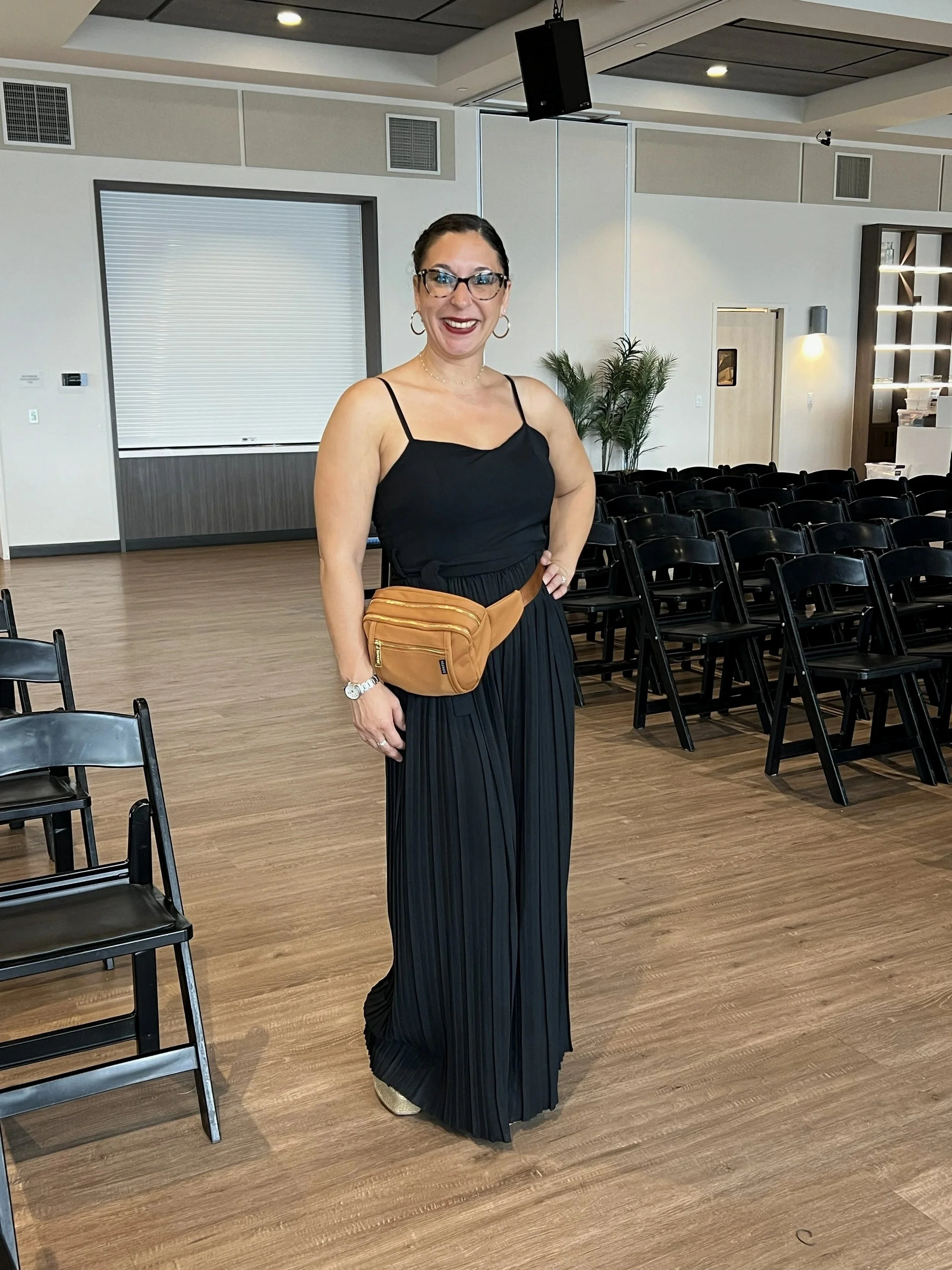 A woman standing in a banquet room with black folding chairs, wearing a black dress with spaghetti straps, glasses, hoop earrings, a watch, and a tan waist bag, smiling at the camera.