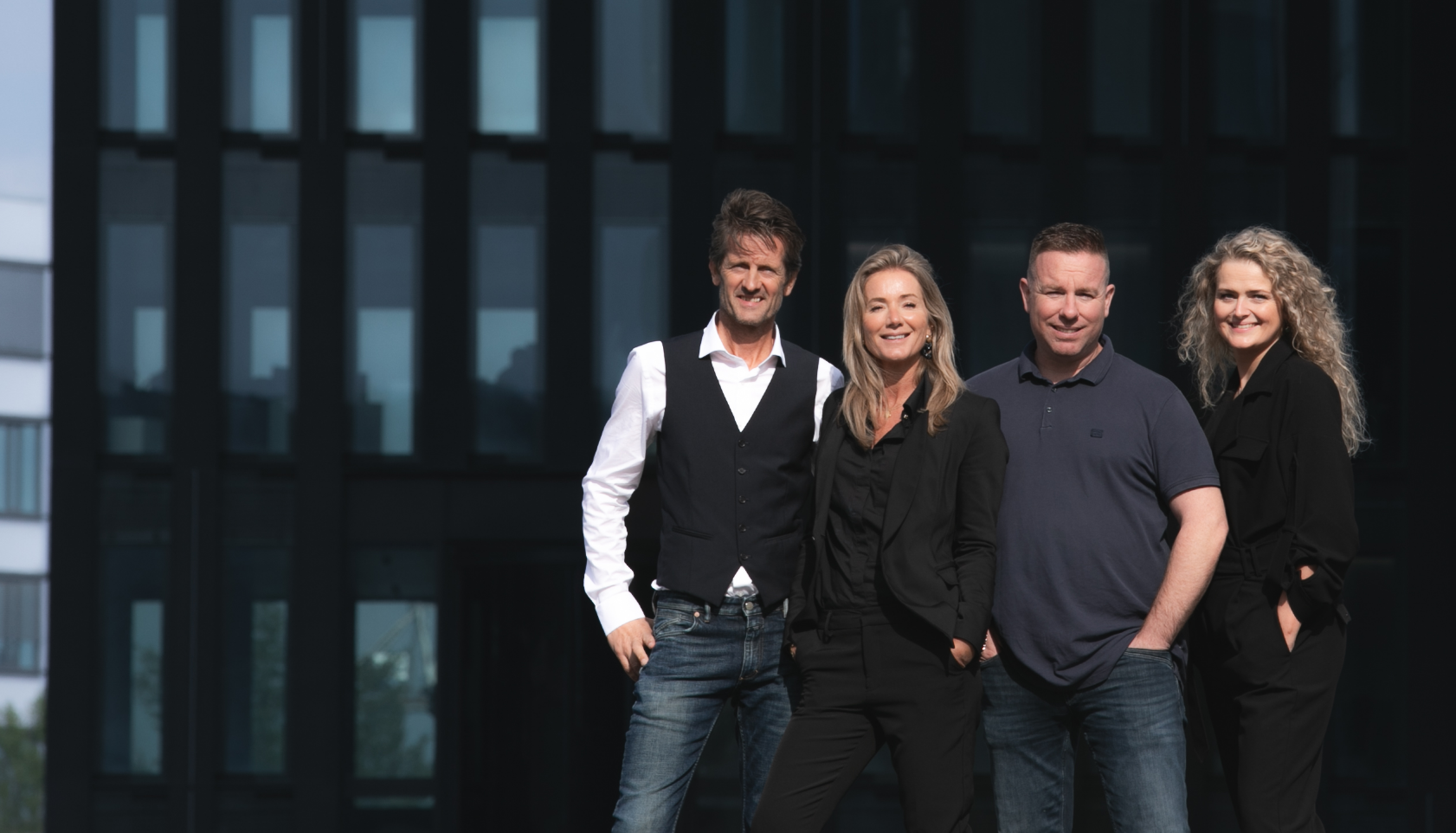 Vijf mensen staan buiten voor een modern gebouw, poseert voor de camera en glimlacht.