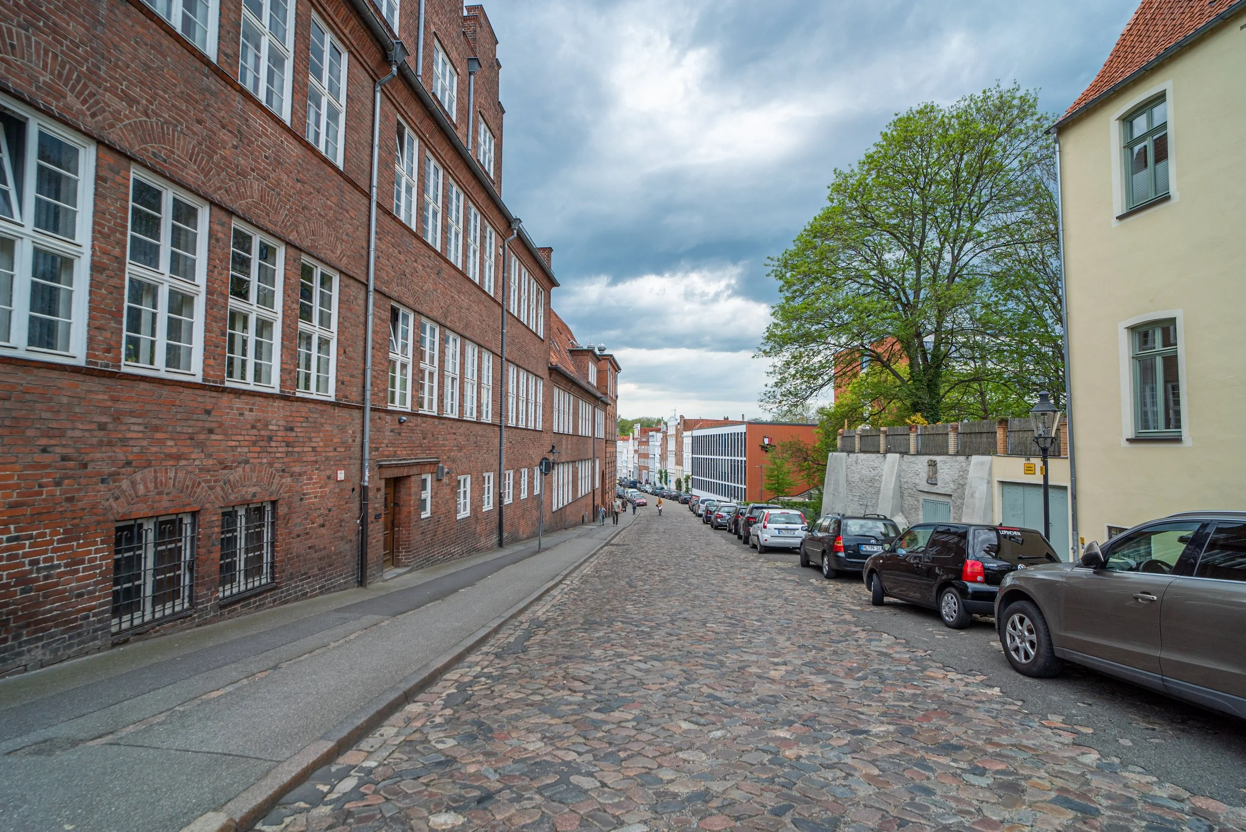 Eine gepflasterte Straße mit an den Seiten geparkten Autos, umgeben von alten und modernen Gebäuden, mit einem großen Baum und einem bewölkten Himmel.