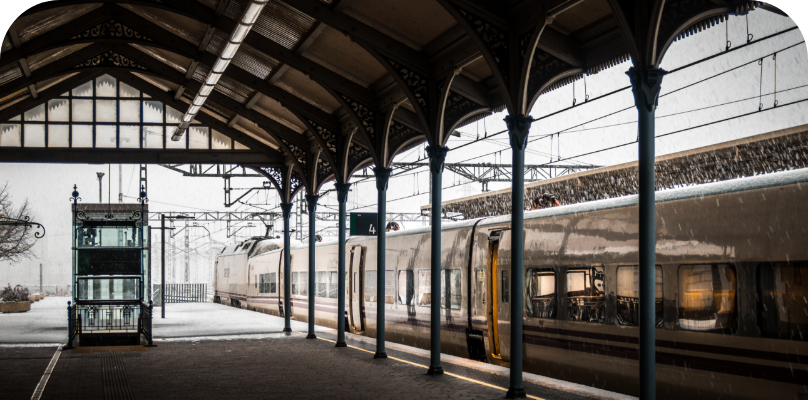 Ein Zug steht still auf einem Bahnsteig unter einem metallenen Bahnhofsdach, der Boden ist mit Schnee bedeckt und der Himmel stark bewölkt.