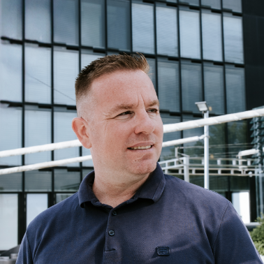 Man met kort bruin haar en een blauwe polo voor een modern kantoorgebouw.