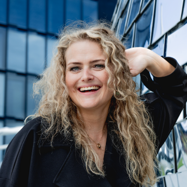 Een lachende vrouw met lang, krullend blond haar poseert buiten, met een modern kantoorpand op de achtergrond.