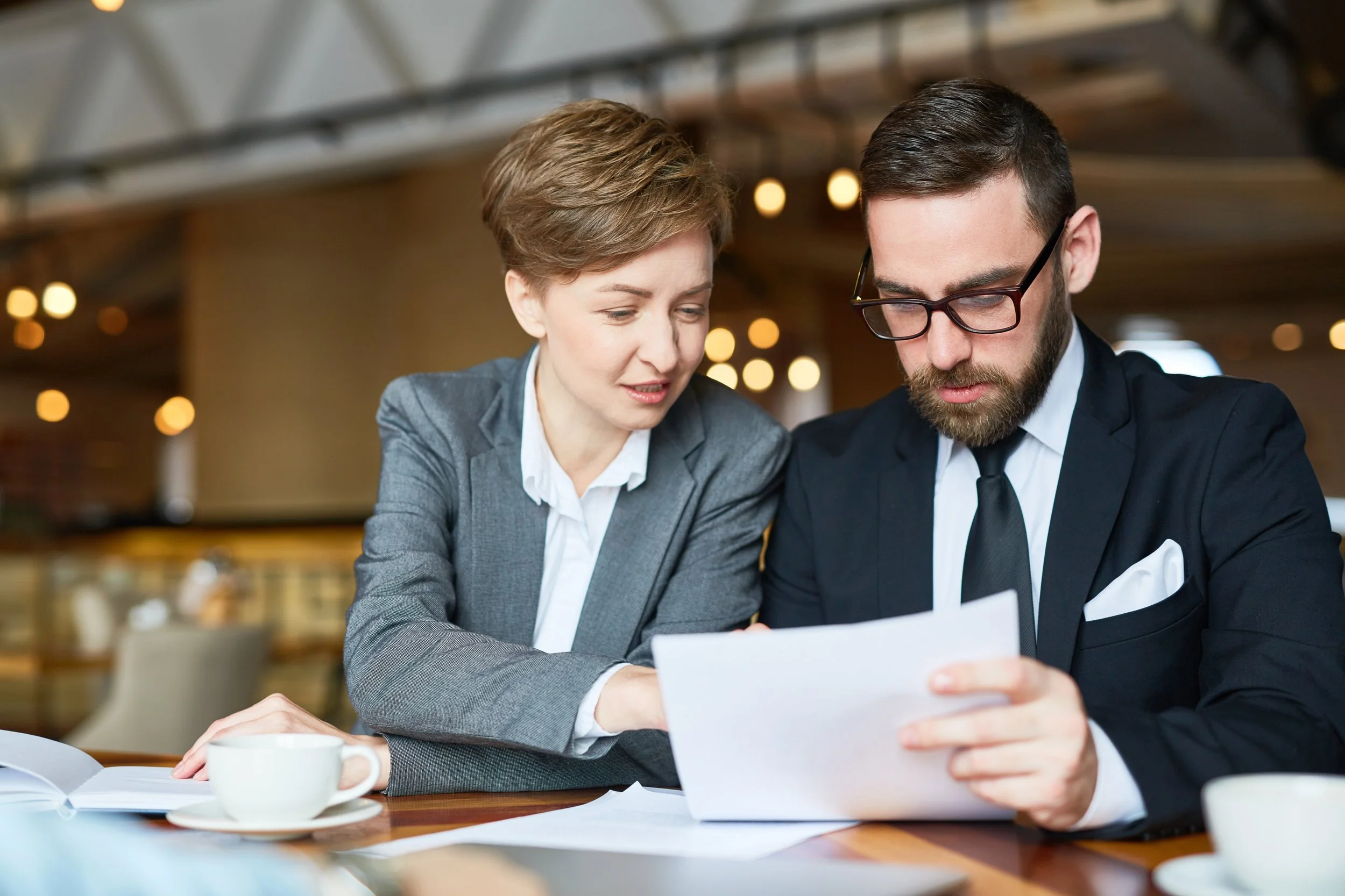 Two professionals, a woman and a man, are sitting at a table looking at a document together in a conference room or cafe setting.