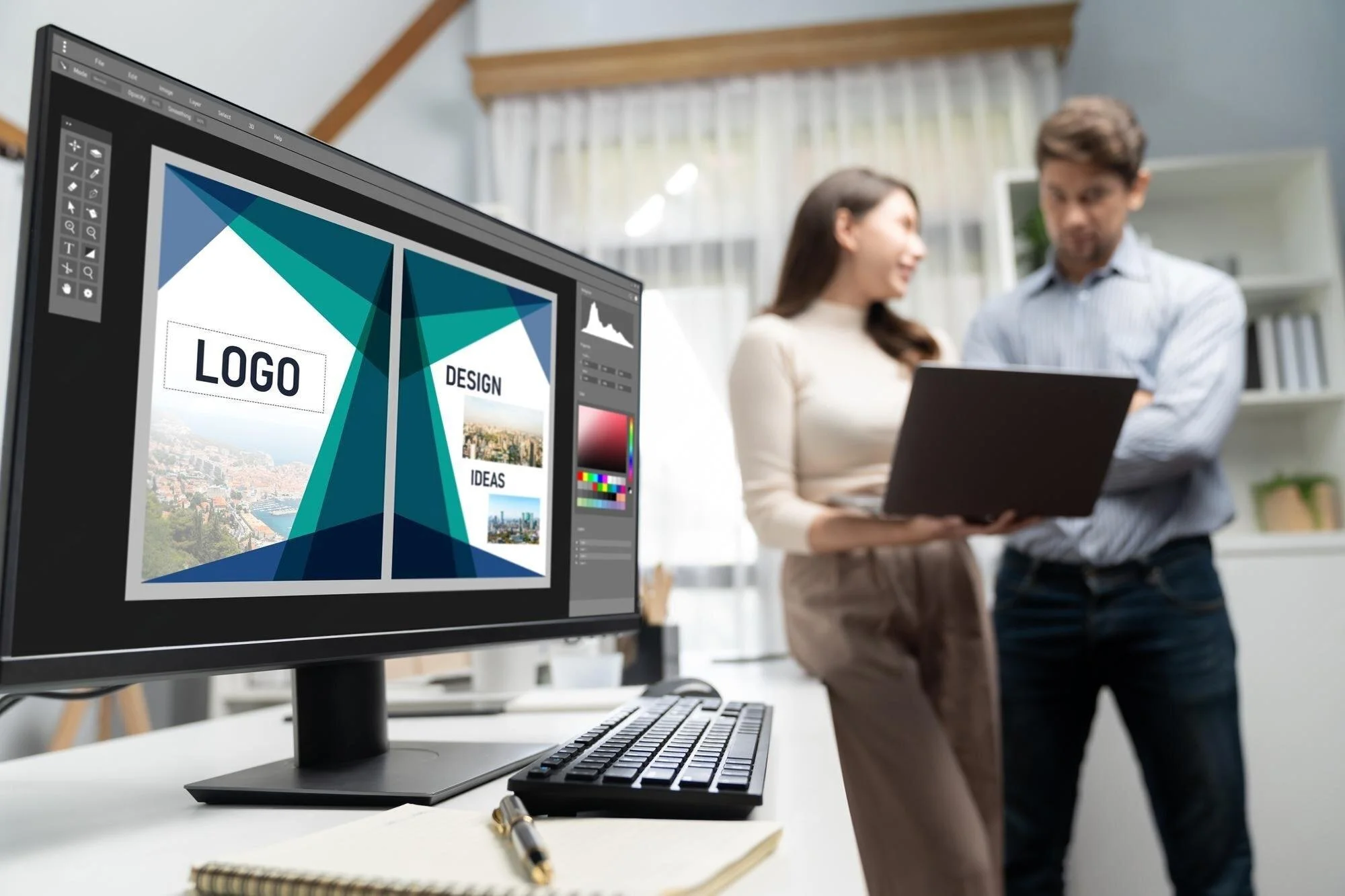 Two people looking at a laptop in an office, with a computer monitor displaying a design project titled 'Logo Design Ideas'.