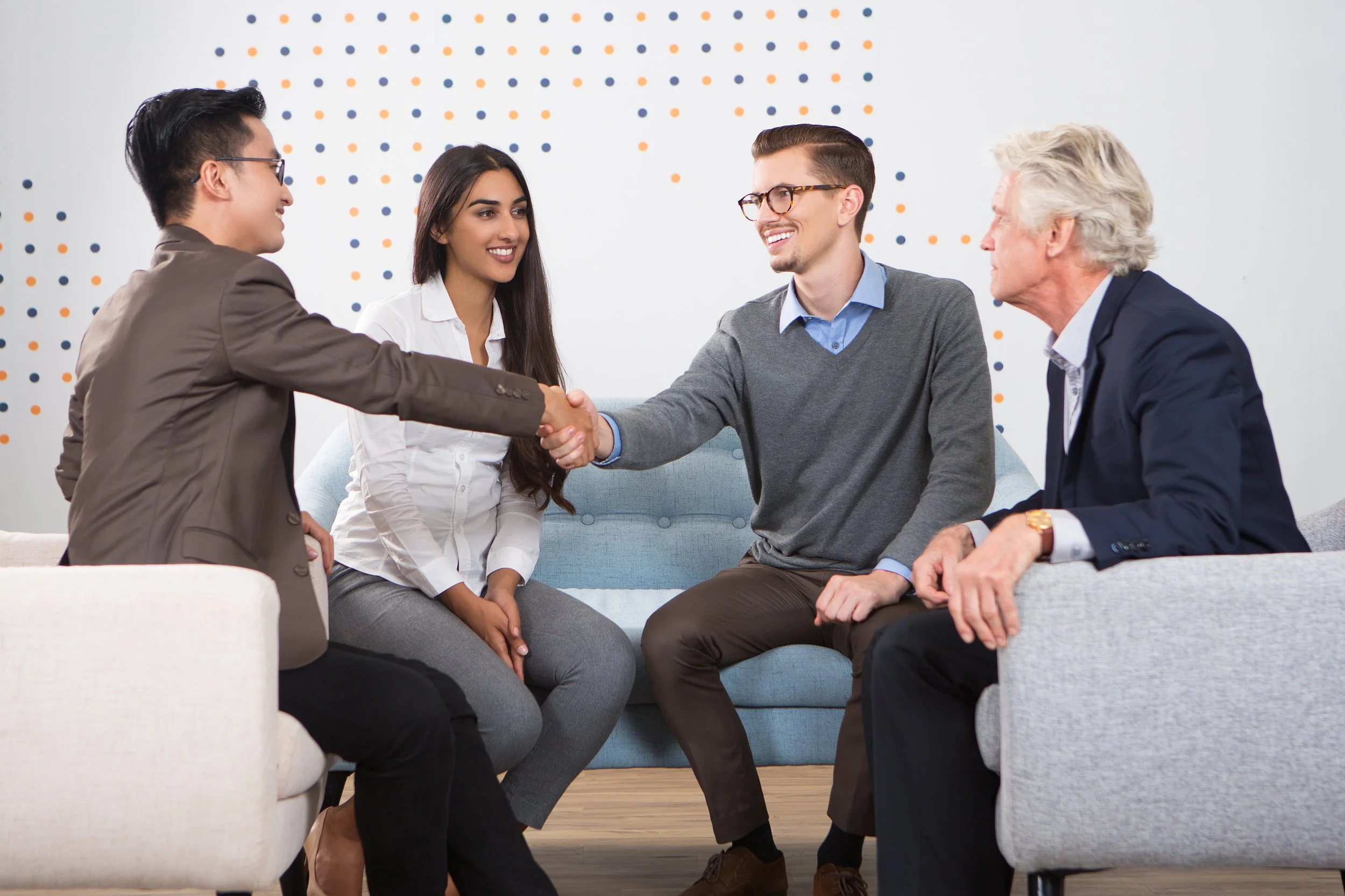 A diverse group of four adults, two men and two women, sitting on couches in an office setting. They are engaged in a friendly meeting, with two of them shaking hands and smiling, indicating a successful business or professional interaction.