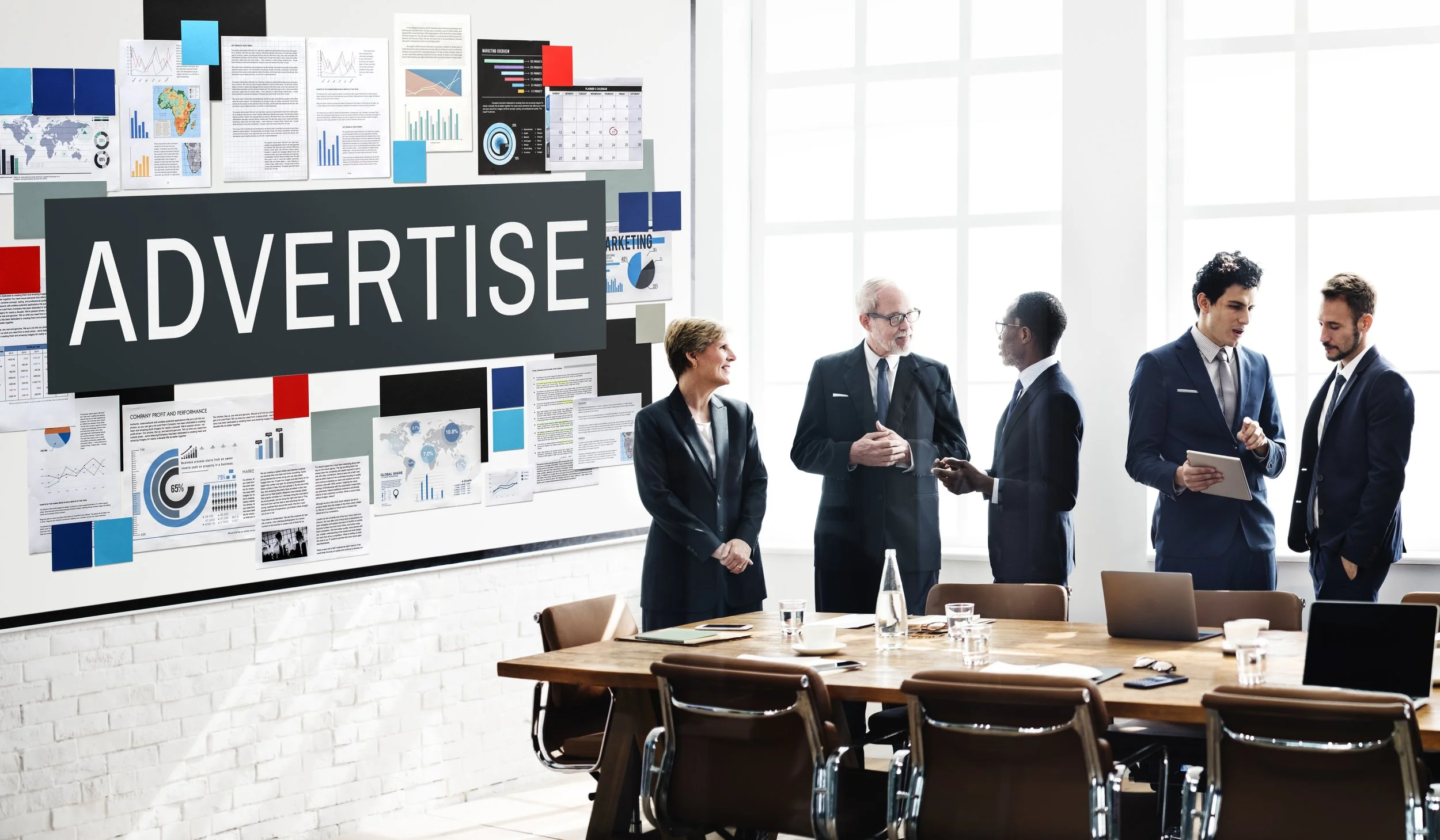 Business meeting in a conference room with a large 'ADVERTISE' display on a wall, six people in suits talking, with tables, laptops, and documents on the table.