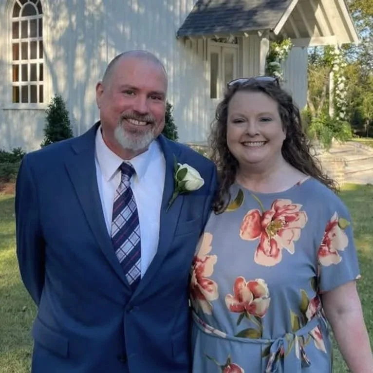A portrait of a man and woman standing outside in front of a white building with a gabled roof. The man is wearing a blue suit, white shirt, and a plaid tie, with a white flower boutonniere. The woman is wearing a light blue floral dress with pink and red flowers and has curly brown hair. Both are smiling.