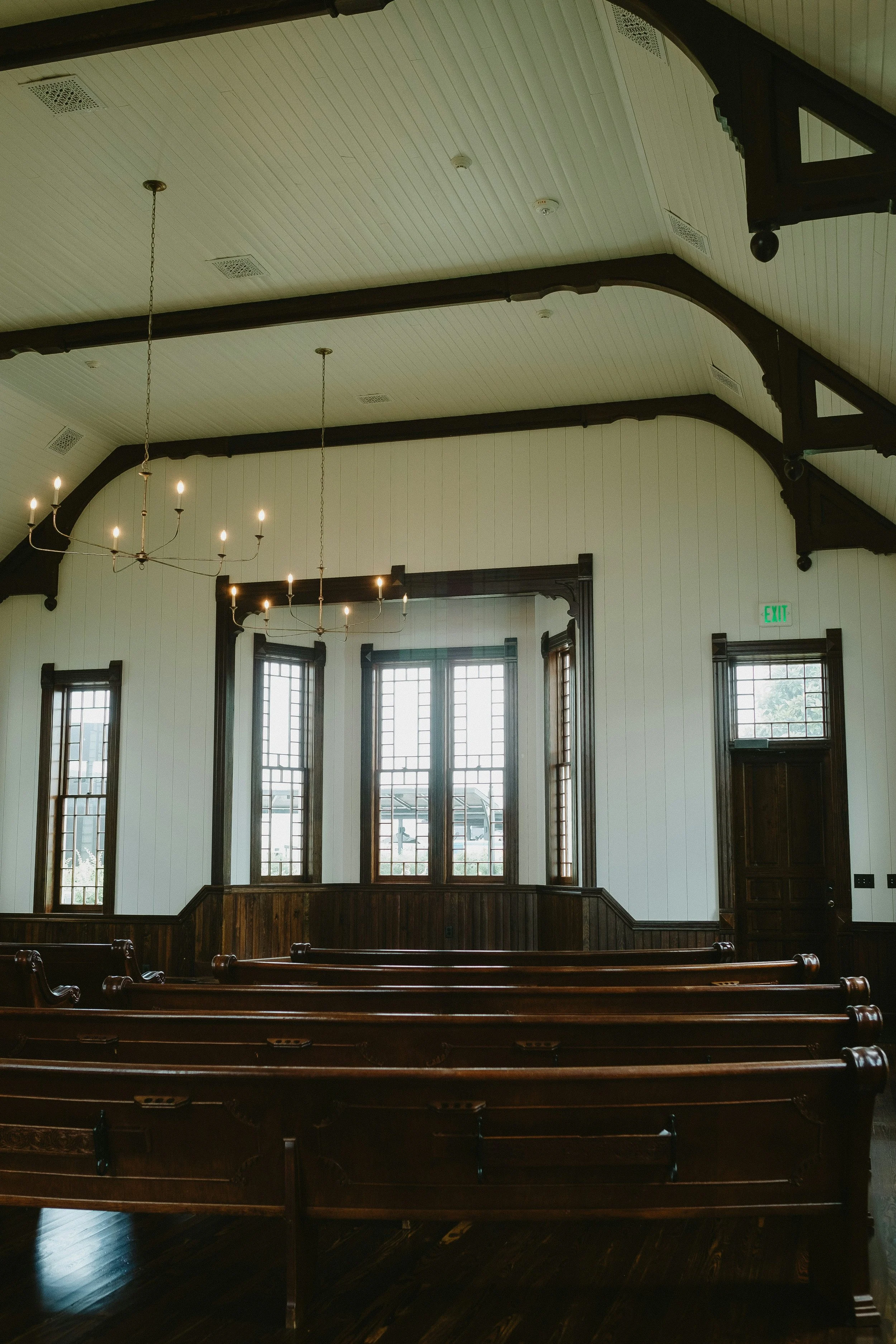 Interior of a church or chapel with wooden pews, tall windows, a chandelier, and a wooden door on the right.
