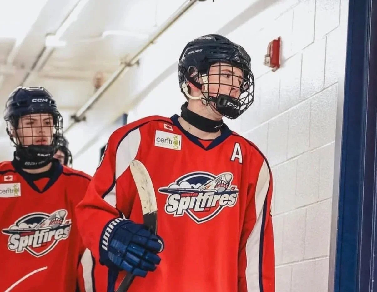 Two ice hockey players in red jerseys and helmets, one with an 'A' on her shoulder, walking in a hockey rink corridor.