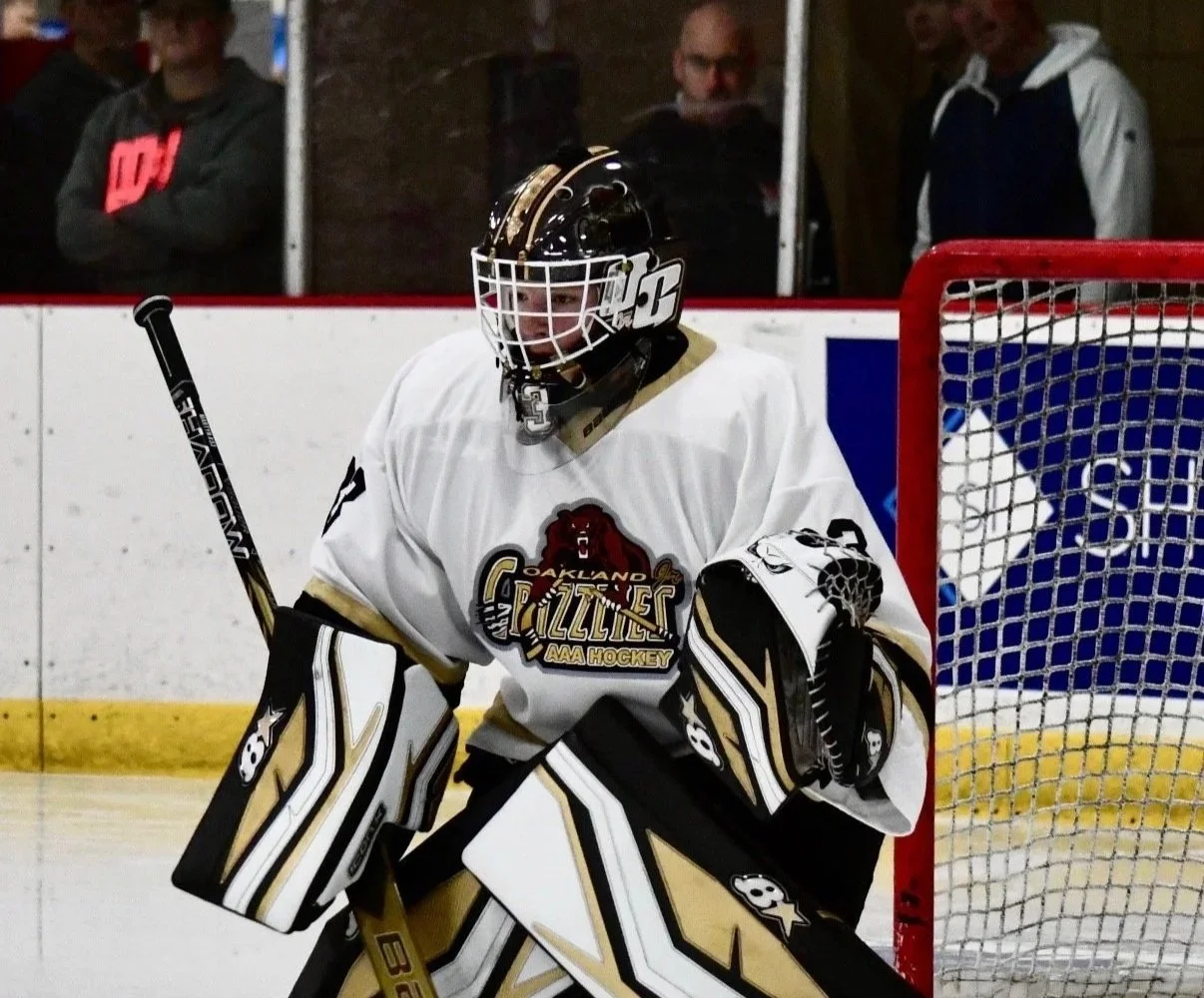 An ice hockey goalie wearing a white jersey with 'Oakland Gazzlies AAA Hockey' logo, black and gold pads, and a black helmet with a face cage, standing in front of the goal net on an ice rink.