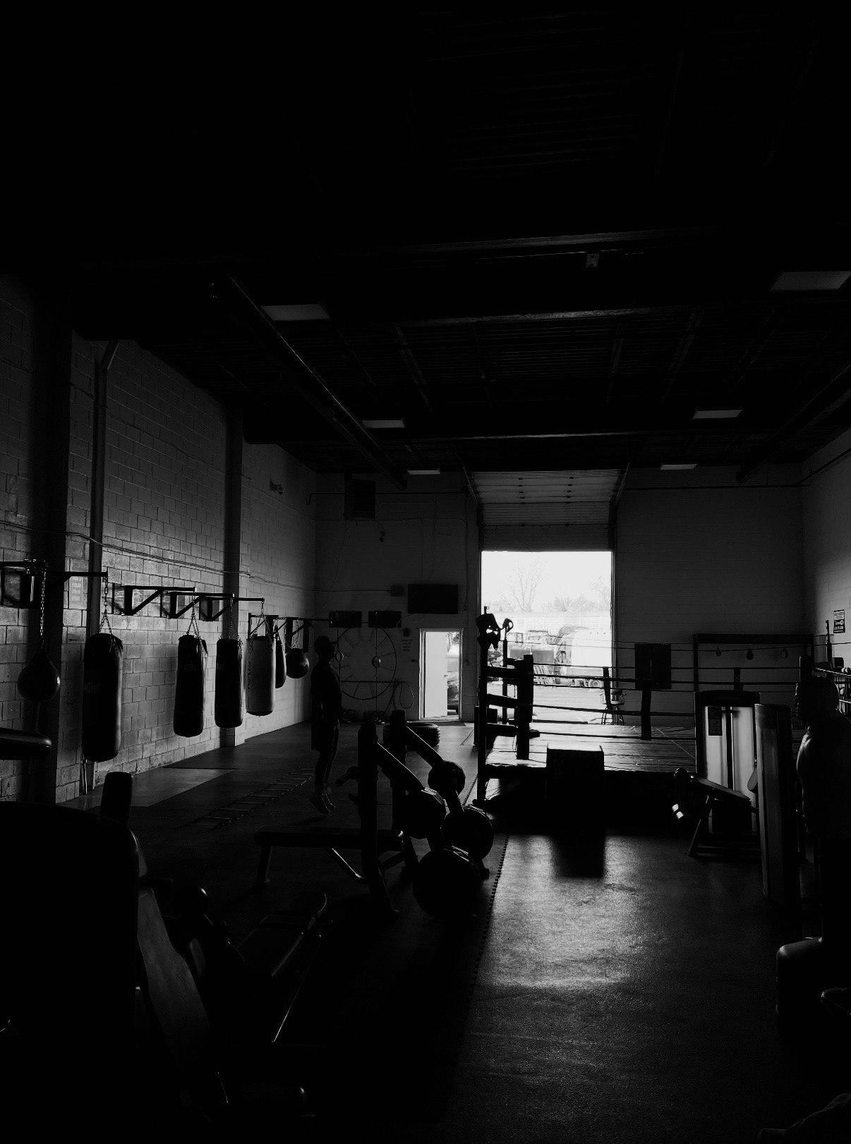 Interior of a boxing gym with hanging punching bags, a boxing ring, and a person standing near the bags, with natural light coming through an open garage door.