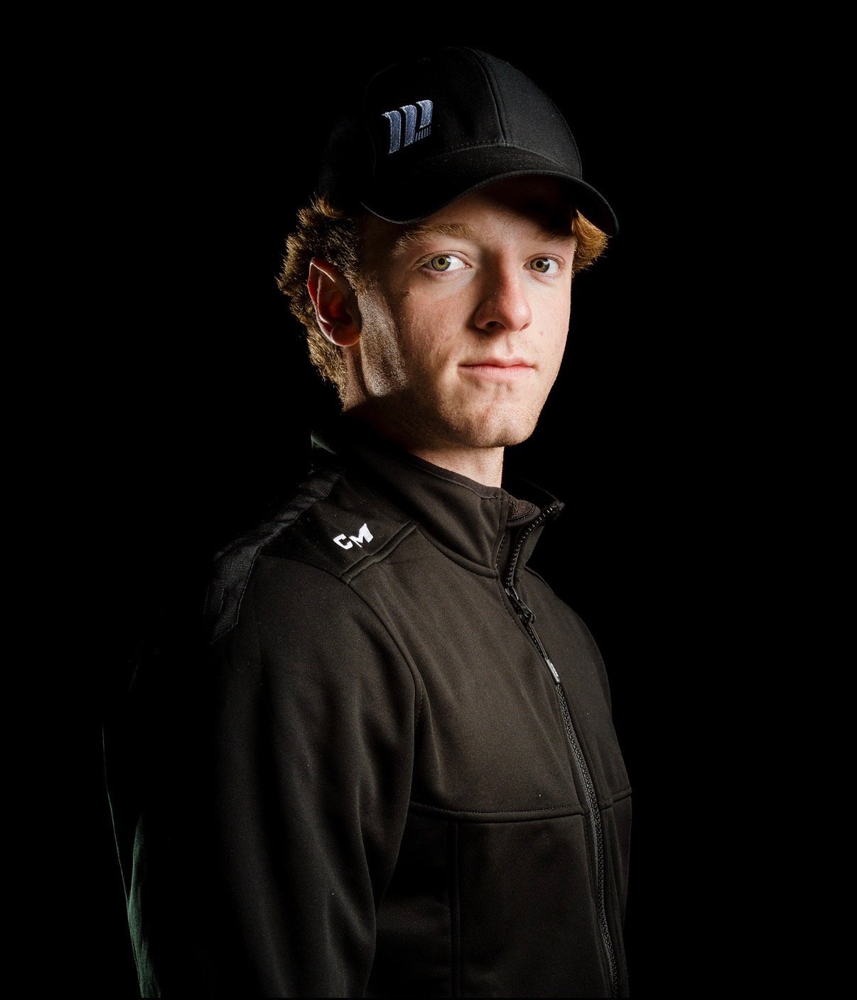 Portrait of a young man wearing a black cap and black athletic jacket, standing against a black background.
