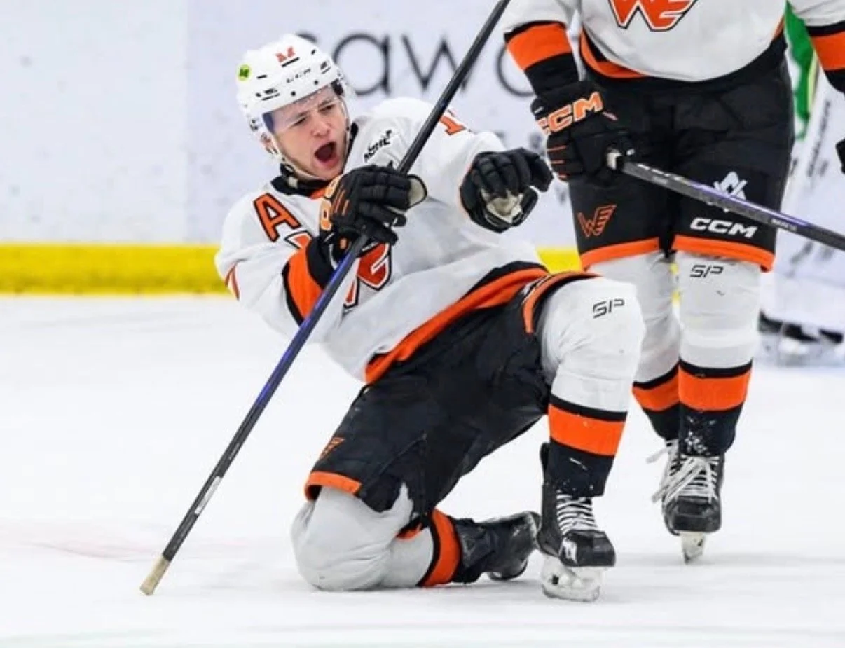 Young male ice hockey player on the ice rink, kneeling on one knee, expressing excitement or celebration, wearing a white and orange jersey, black hockey pants, and holding a hockey stick.