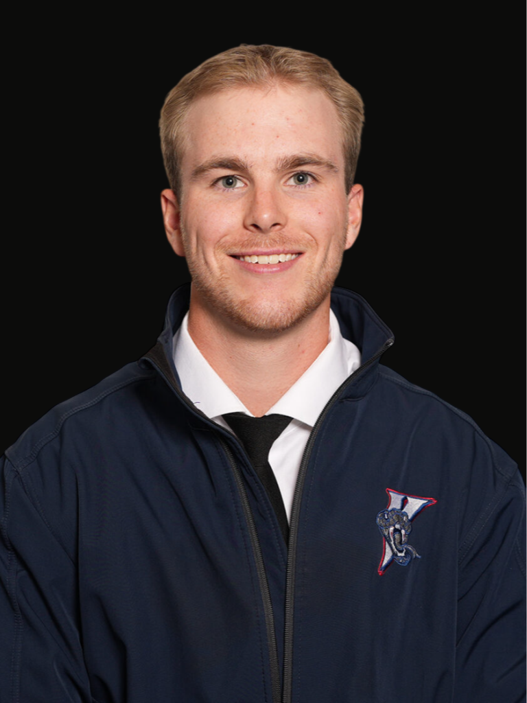 Portrait of a young man with short blond hair and blue eyes, wearing a navy blue jacket with a sports team's logo on the chest, a white shirt, and a black tie, smiling against a black background.