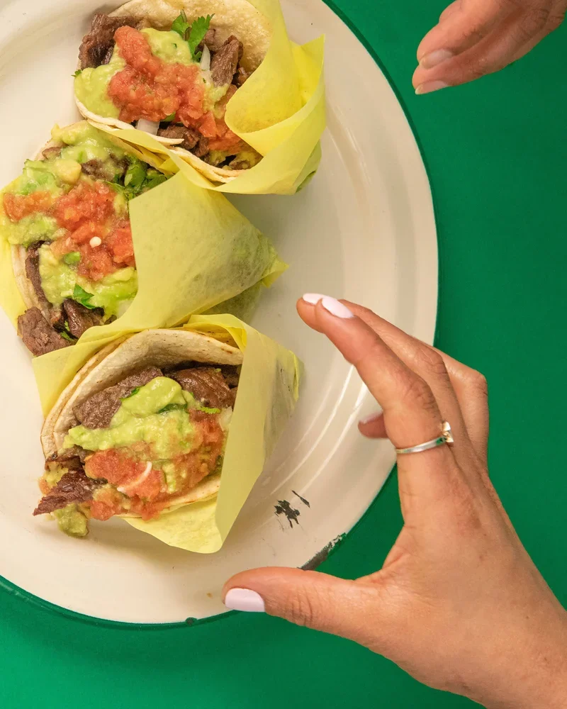 Three tacos filled with beef, guacamole, and tomato, wrapped in yellow paper on a white plate with a green background.