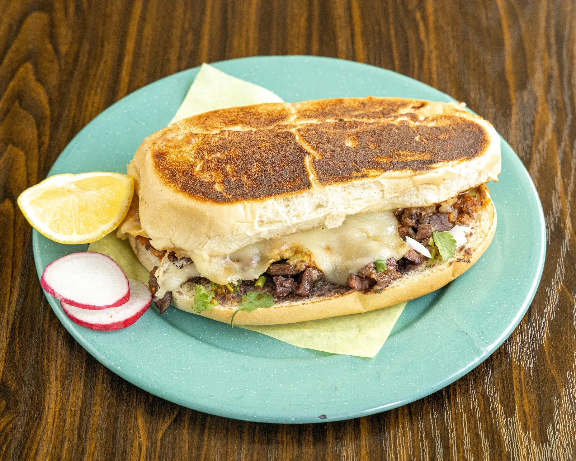 A hoagie sandwich on a blue plate with melted cheese, beef and vegetables, served with lemon wedge and radish slices, on a wooden table.
