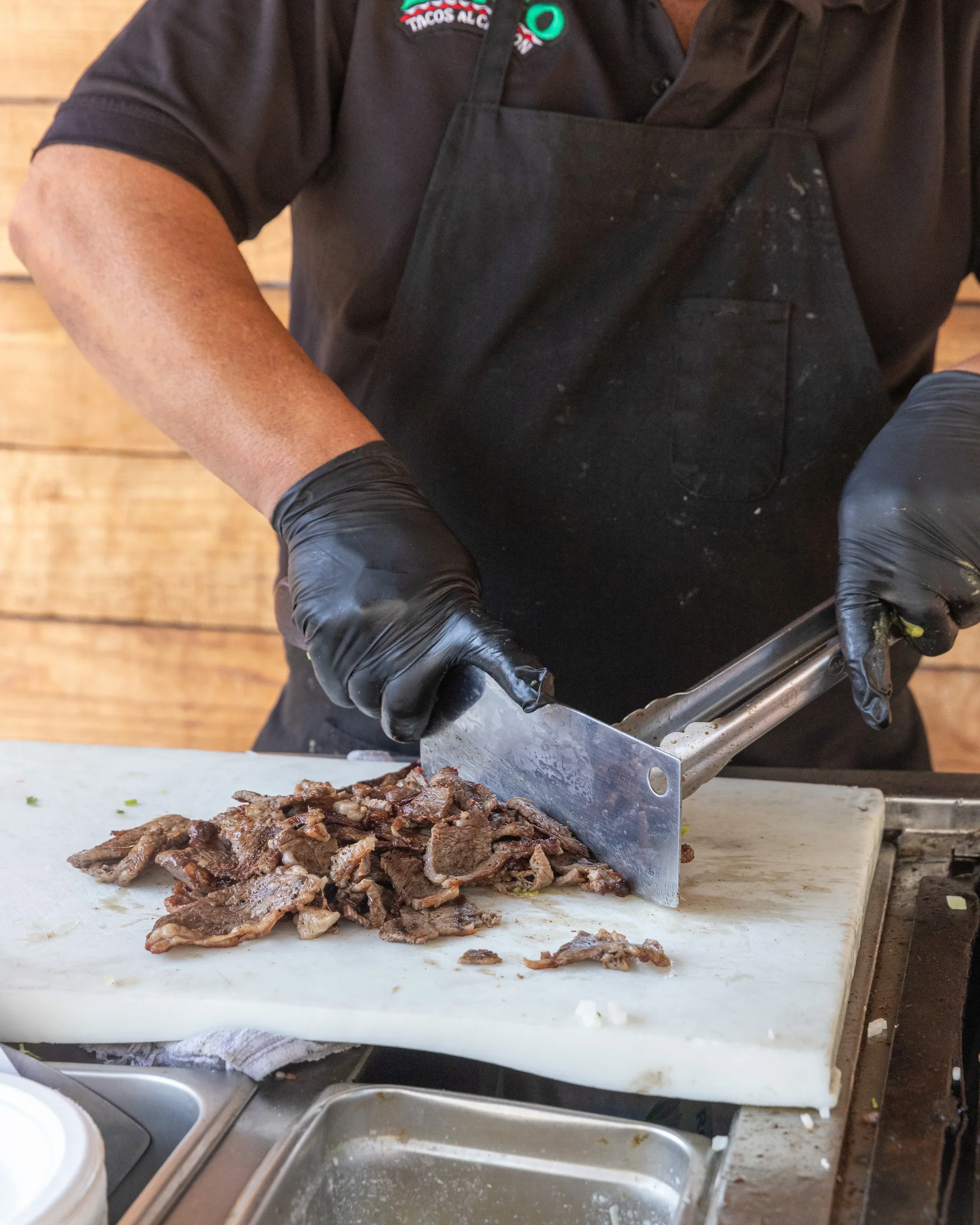 Person in black apron and gloves chopping cooked meat on a white cutting board.