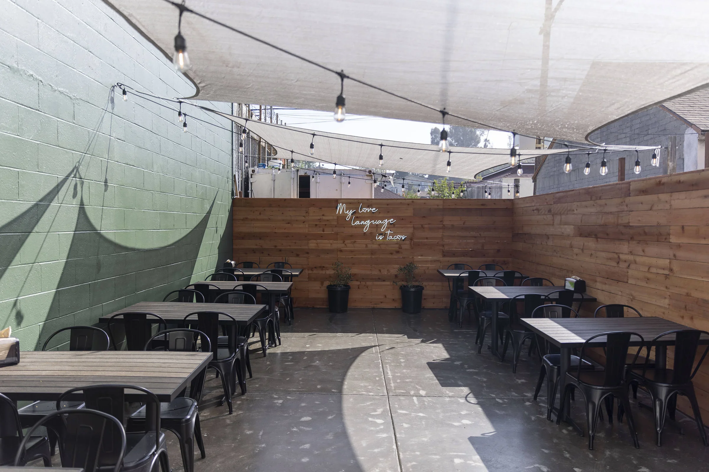 Outdoor dining area with black tables and chairs, string lights overhead, a green wall on the left, and a wood fence on the back wall with a neon sign that reads 'My Love Language is Tacos'.