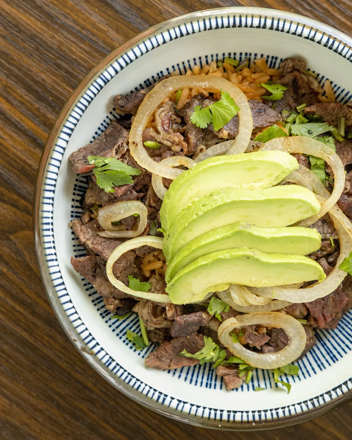 A bowl of carne asada topped with sliced onions, avocado slices, and fresh cilantro, served on a wooden table.