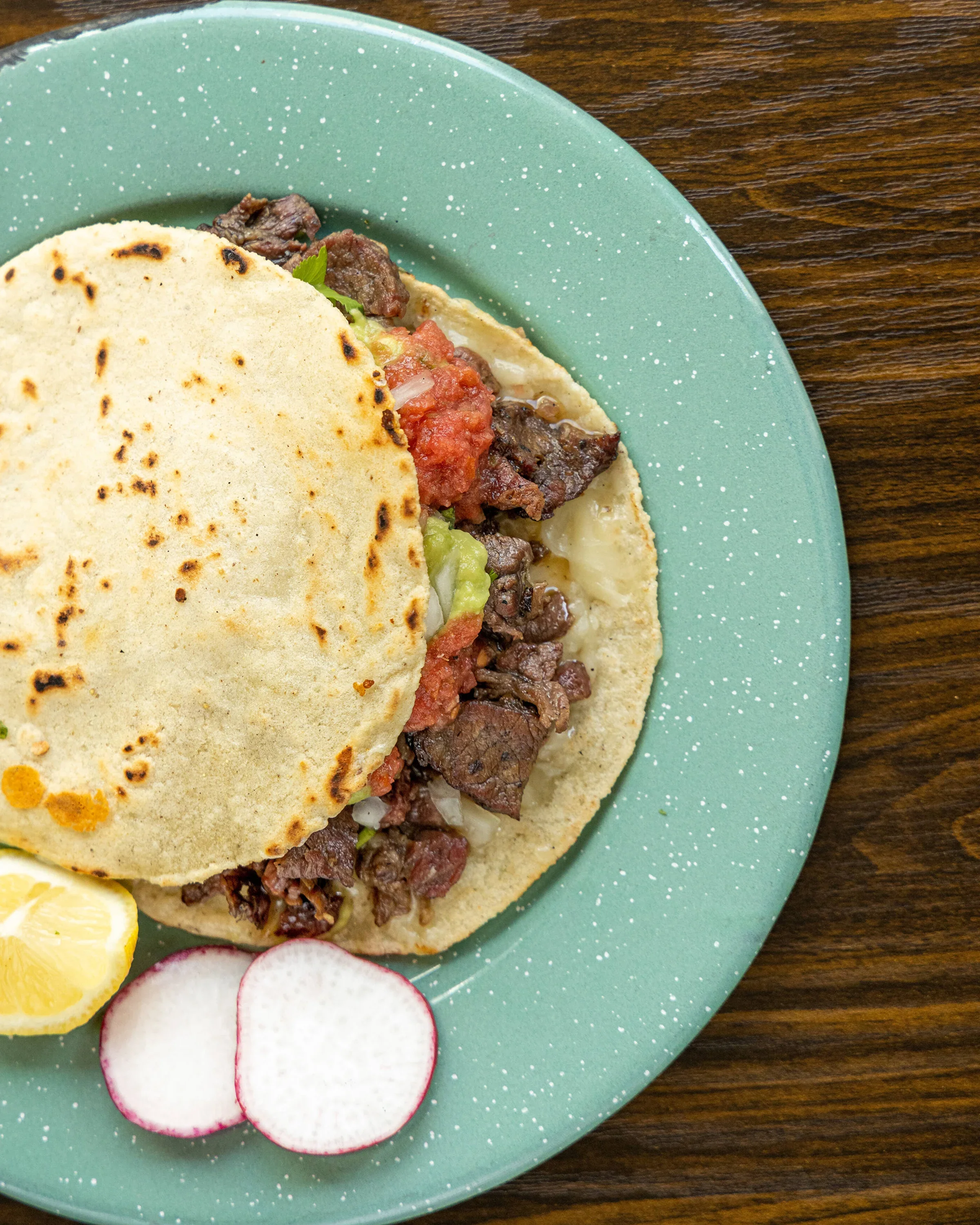 Close-up of a beef taco with rice, tomatoes, lettuce, radishes, and lemon wedges on a green plate.
