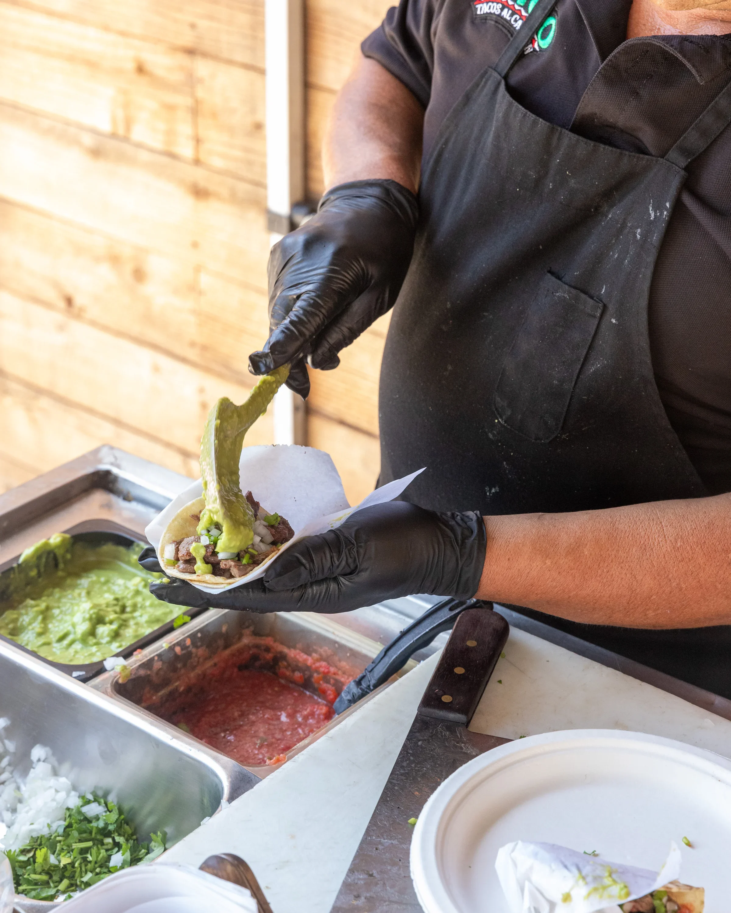 Person assembling a taco with guacamole, salsa, and toppings at a food stand.