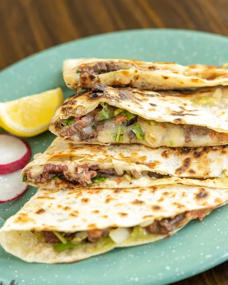 Four beef and cheese quesadillas served on a teal plate, garnished with lemon wedges and sliced radishes.