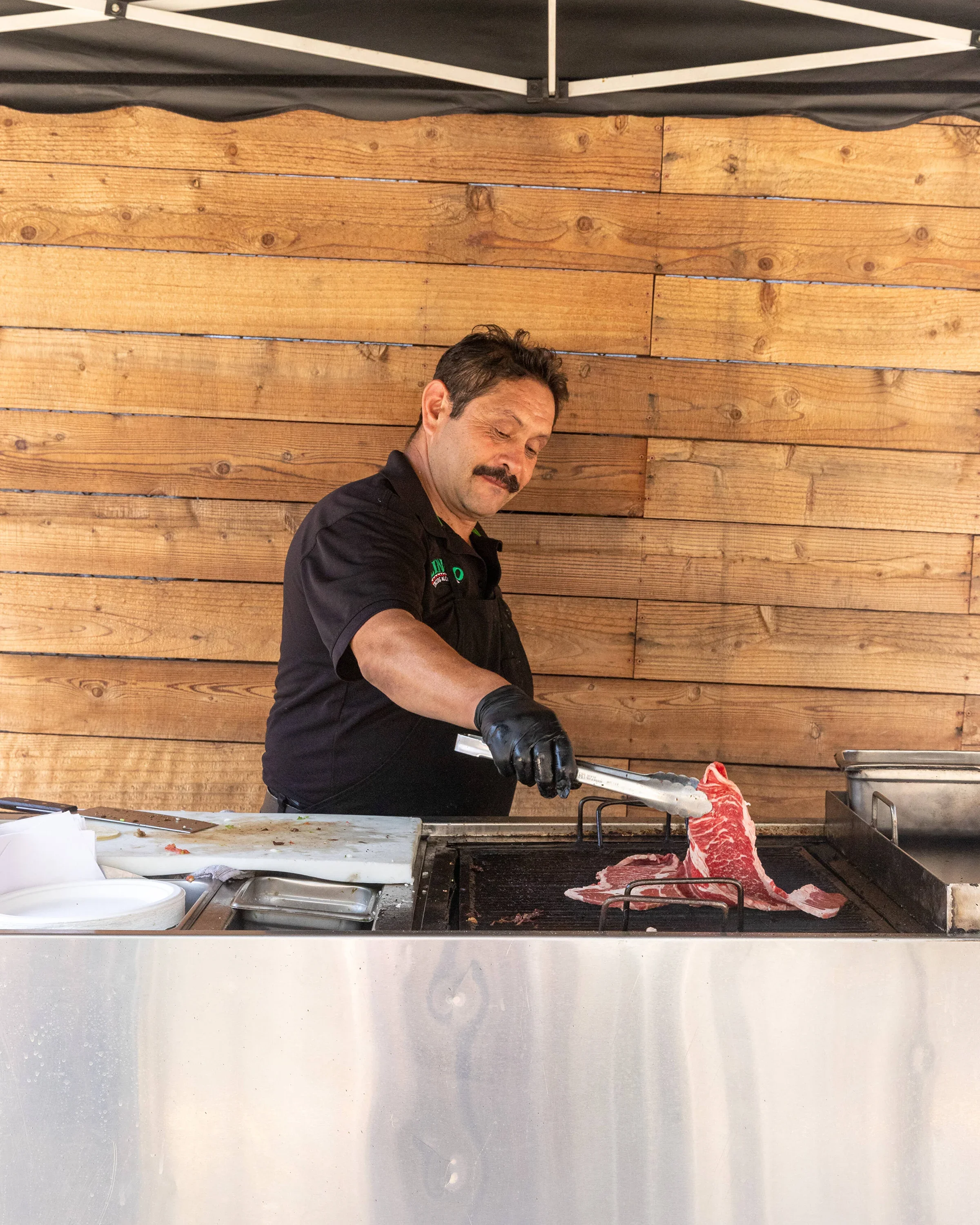 A man grilling a piece of marbled beef on a barbecue grill, wearing black gloves and a black shirt, with a wooden wall in the background.